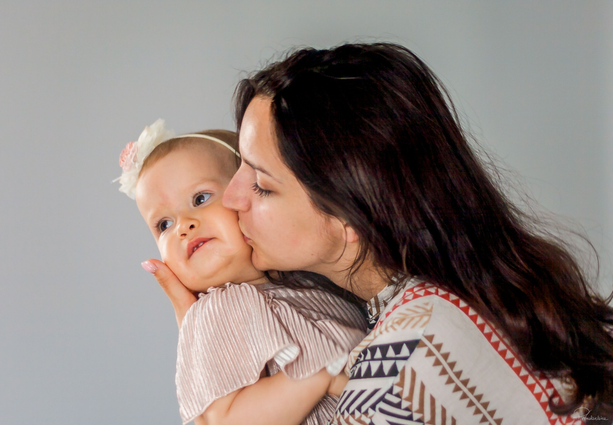 Mama całuje w policzek roczną dziewczynkę z kwiatkiem we włosach, na jasnym tle. Uśmiechnięte dziecko patrzy w bok. Ujęcie z bliska, ciepłe światło.