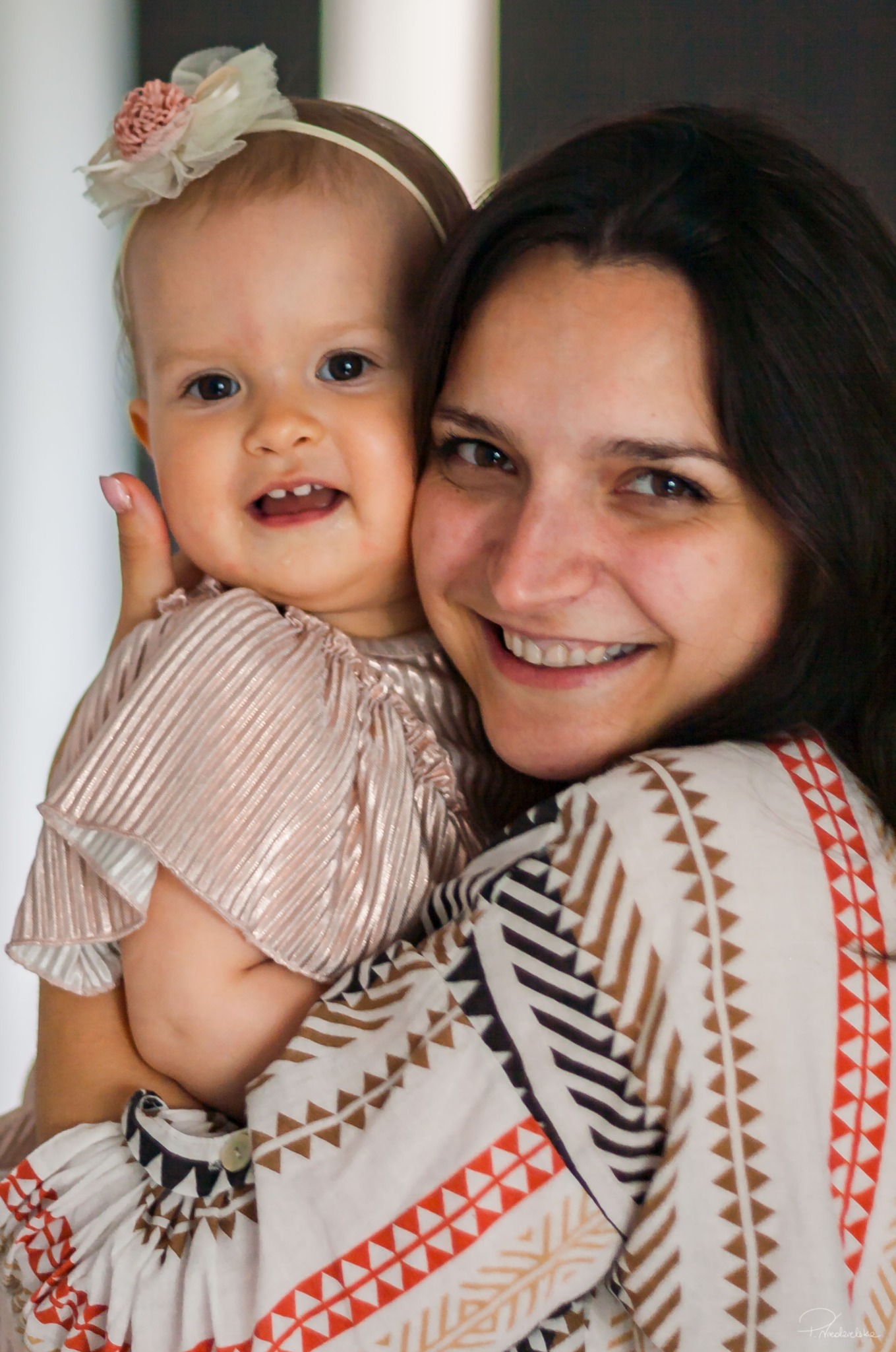 Uśmiechnięta mama tuli roczną córeczkę z opaską i w błyszczącej sukience. Kobieta w bluzce z etnicznym wzorem. Zdjęcie portretowe w jasnym oświetleniu.