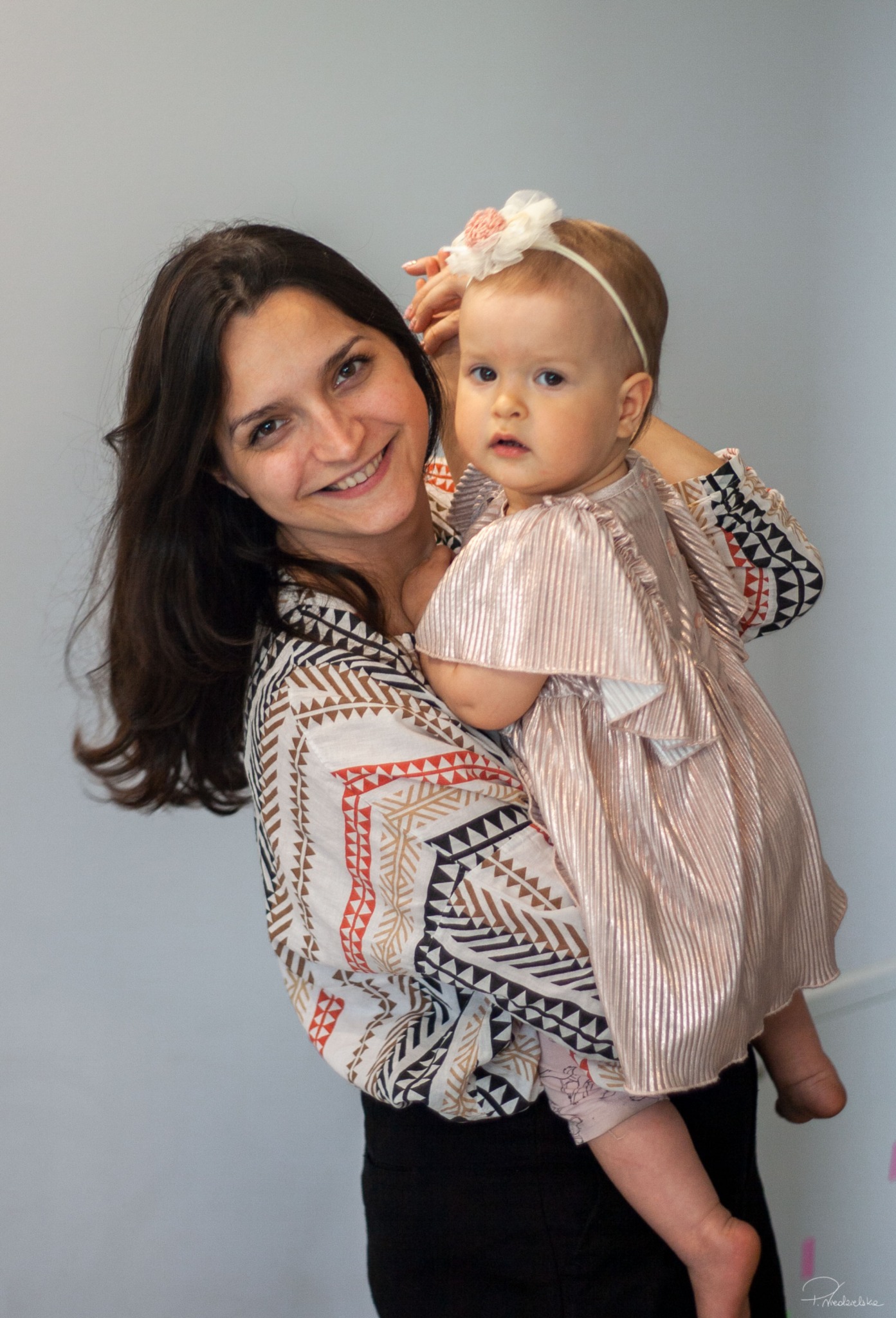 Uśmiechnięta kobieta trzyma na rękach dziewczynkę w błyszczącej sukience i opasce z kwiatkiem, na jasnym tle.