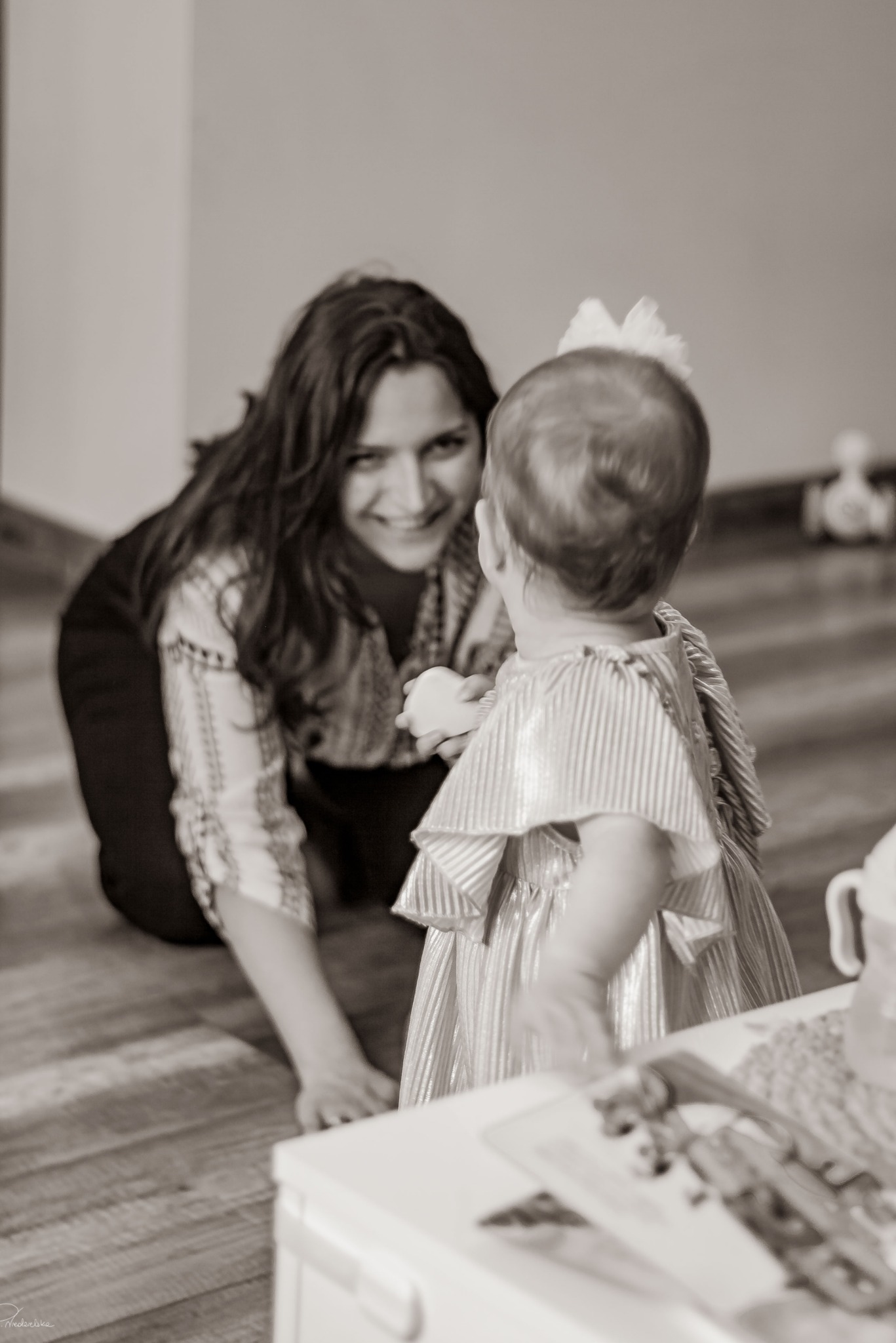 Uśmiechnięta kobieta w bluzce z haftem patrzy na małą dziewczynkę w sukience w paski. Sepia, ciepłe światło, drewniana podłoga.