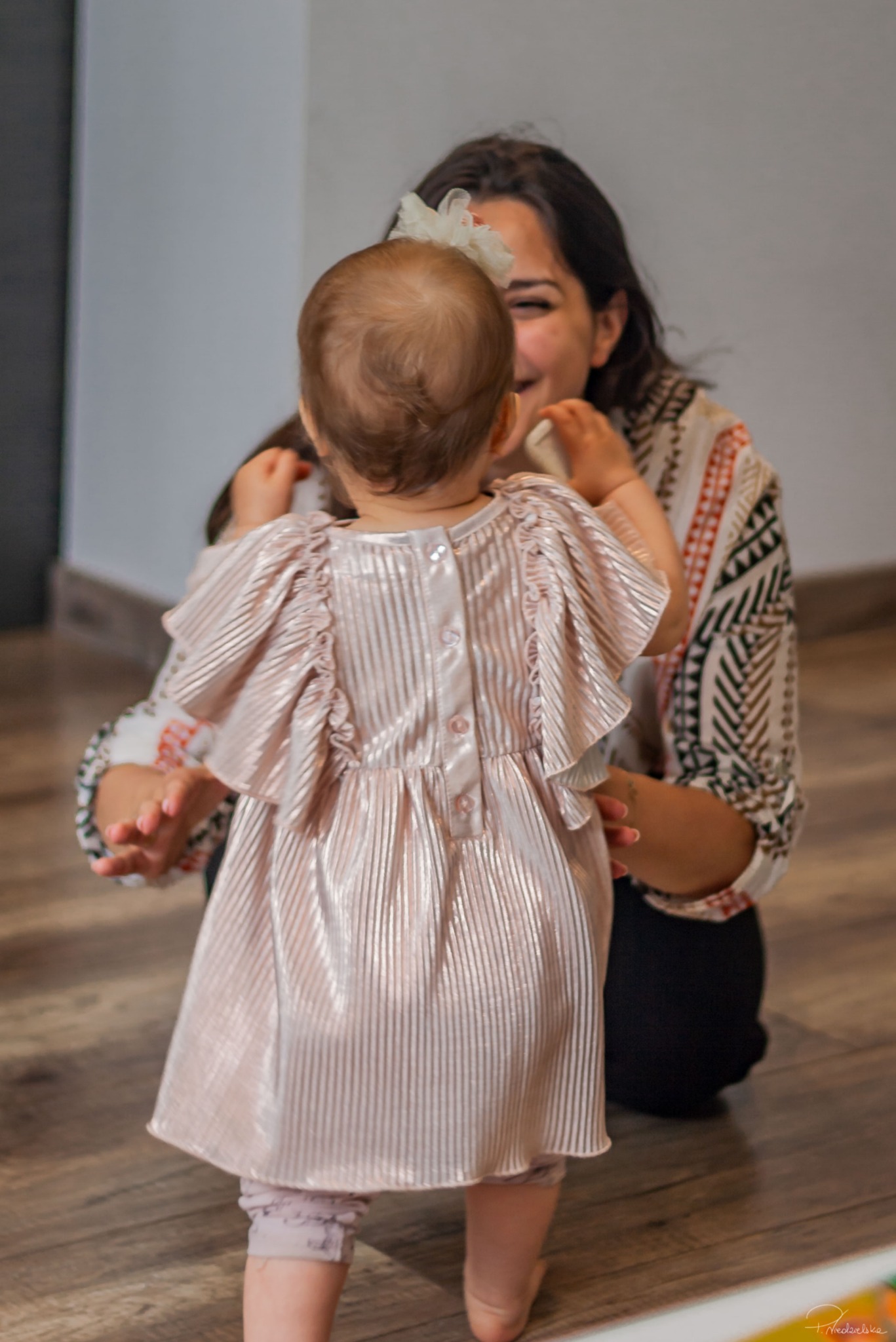 Mama uśmiecha się do rocznej córki w błyszczącej sukience. Dziewczynka stoi tyłem, widać kokardę we włosach i plecy. Jasne, naturalne światło.