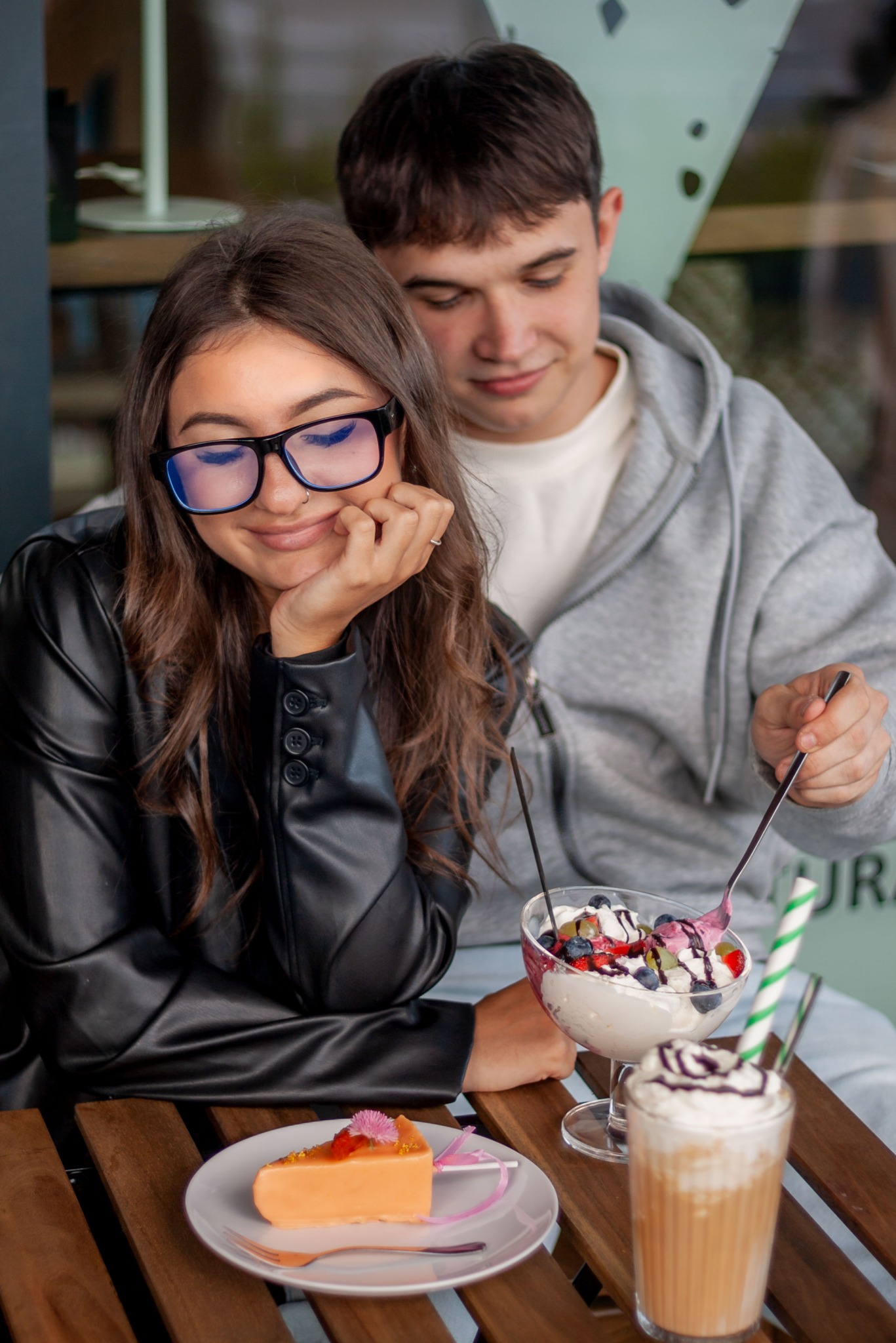 Uśmiechnięta kobieta w okularach i skórzanej kurtce siedzi z mężczyzną przy stoliku z deserami: ciastem, lodami i kawą, w jasnym oświetleniu kawiarni.