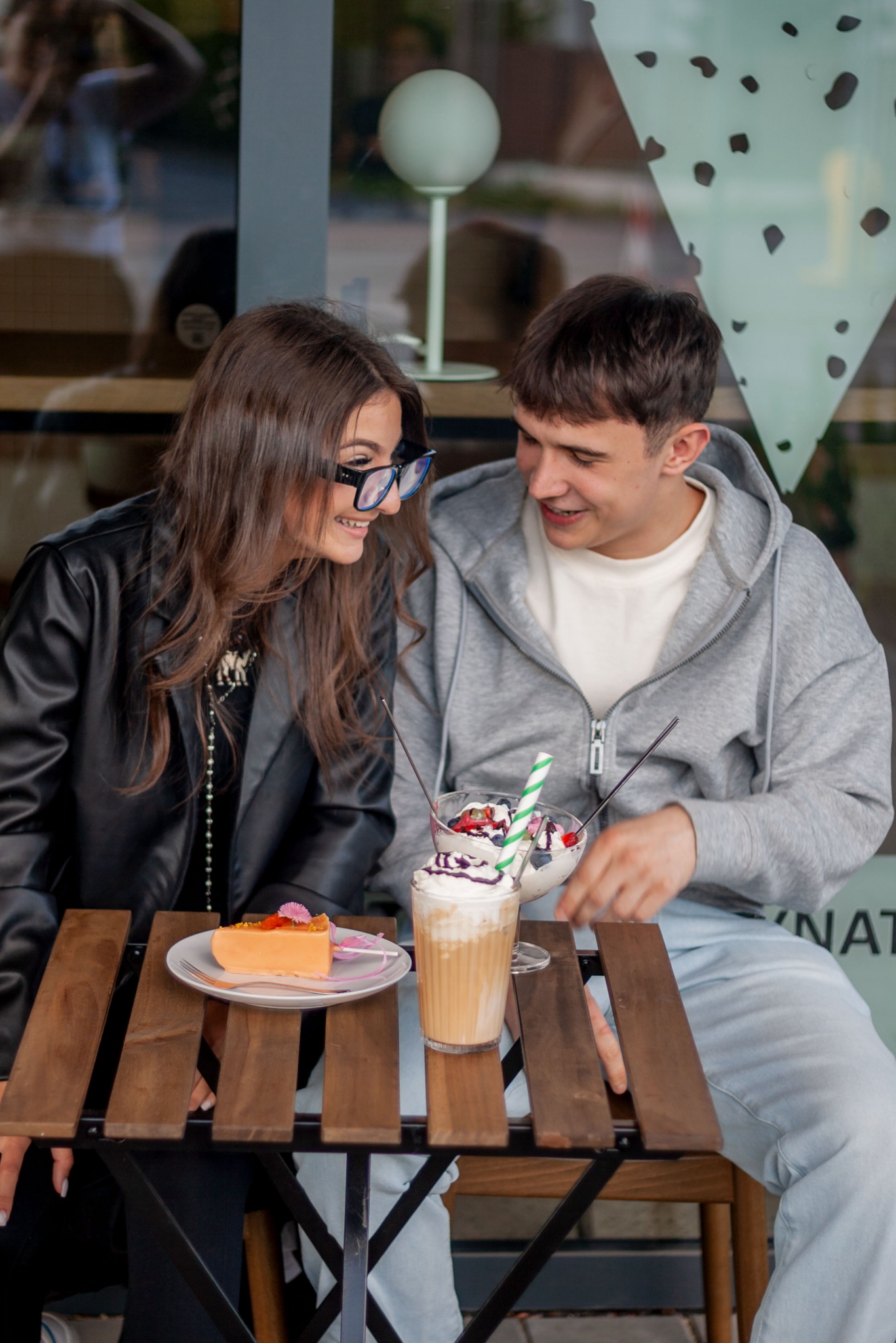 Uśmiechnięta para siedzi przy stoliku w kawiarni, delektując się deserami i napojami. Dziewczyna w okularach i chłopak w bluzie spoglądają na siebie.