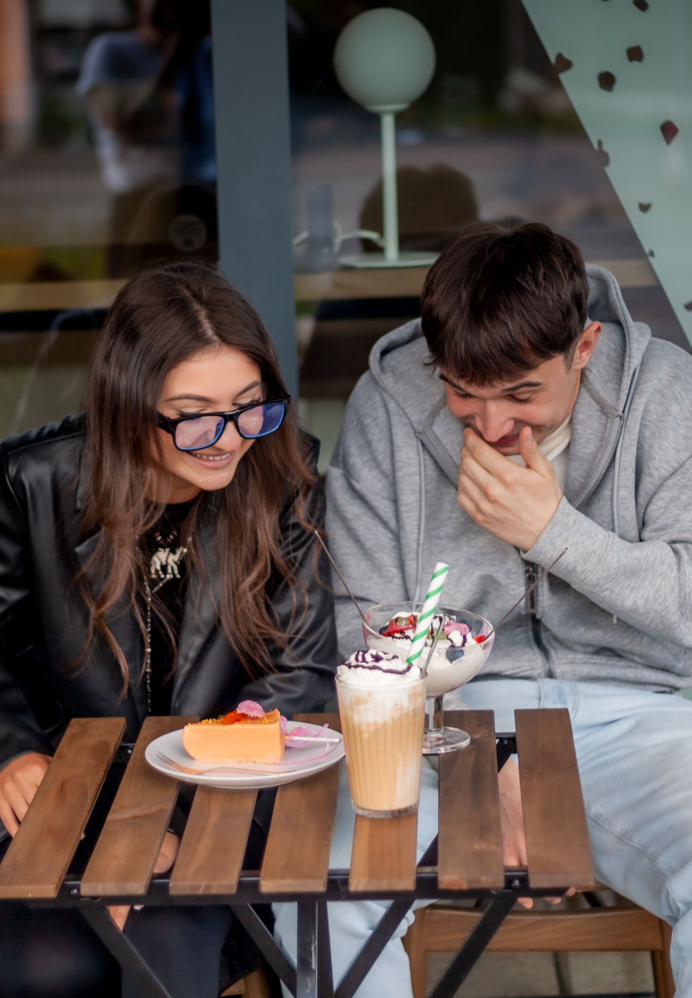Uśmiechnięta para siedzi przy drewnianym stoliku w kawiarni, zajadając deser i pijąc kawę. Dziewczyna w okularach i chłopak w bluzie z kapturem.