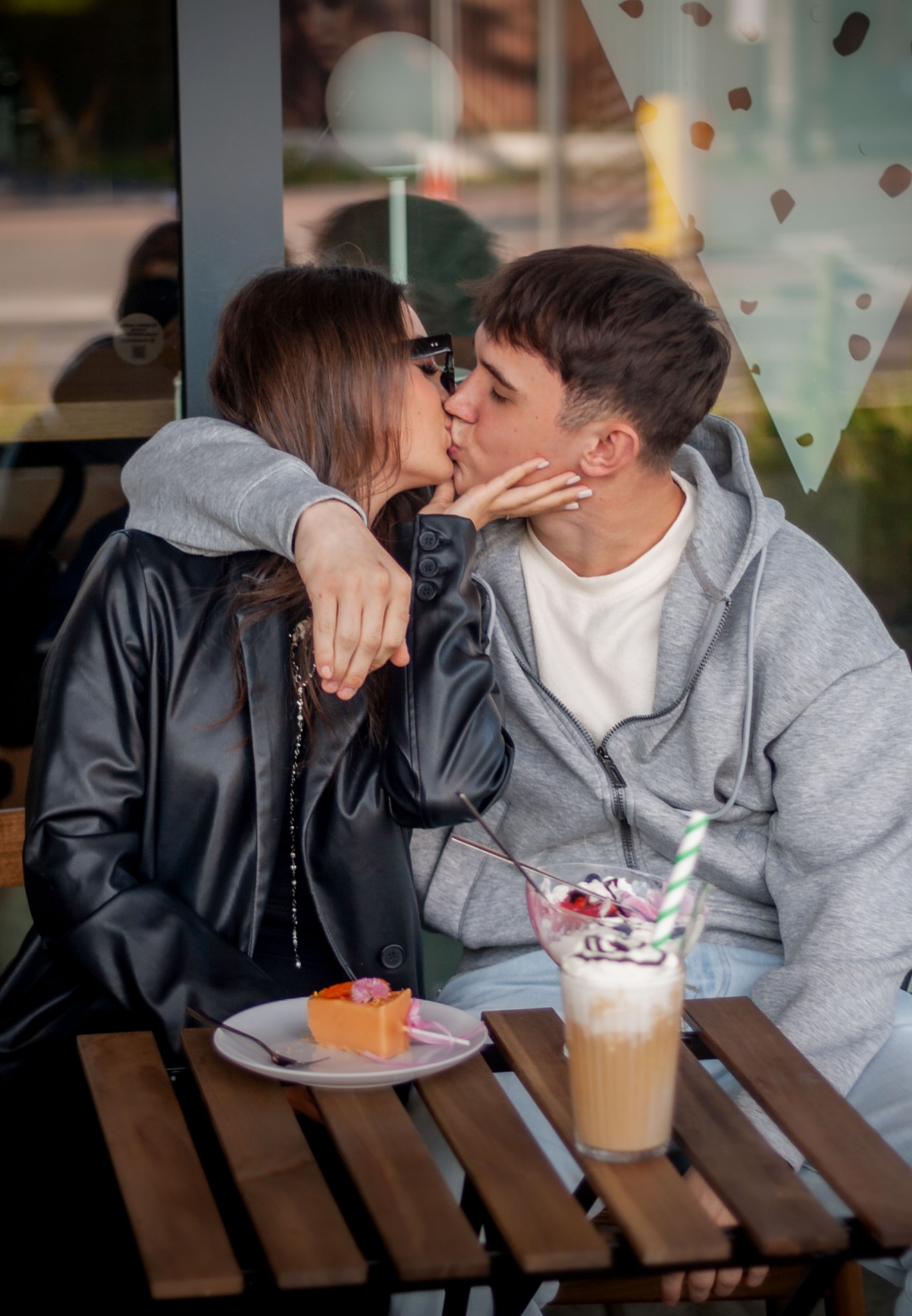 Zakochana para całuje się przy stoliku kawiarnianym, obok desery i napoje. Ona w czarnej kurtce, on w szarej bluzie. Ujęcie z bliska, na pierwszym planie jedzenie.