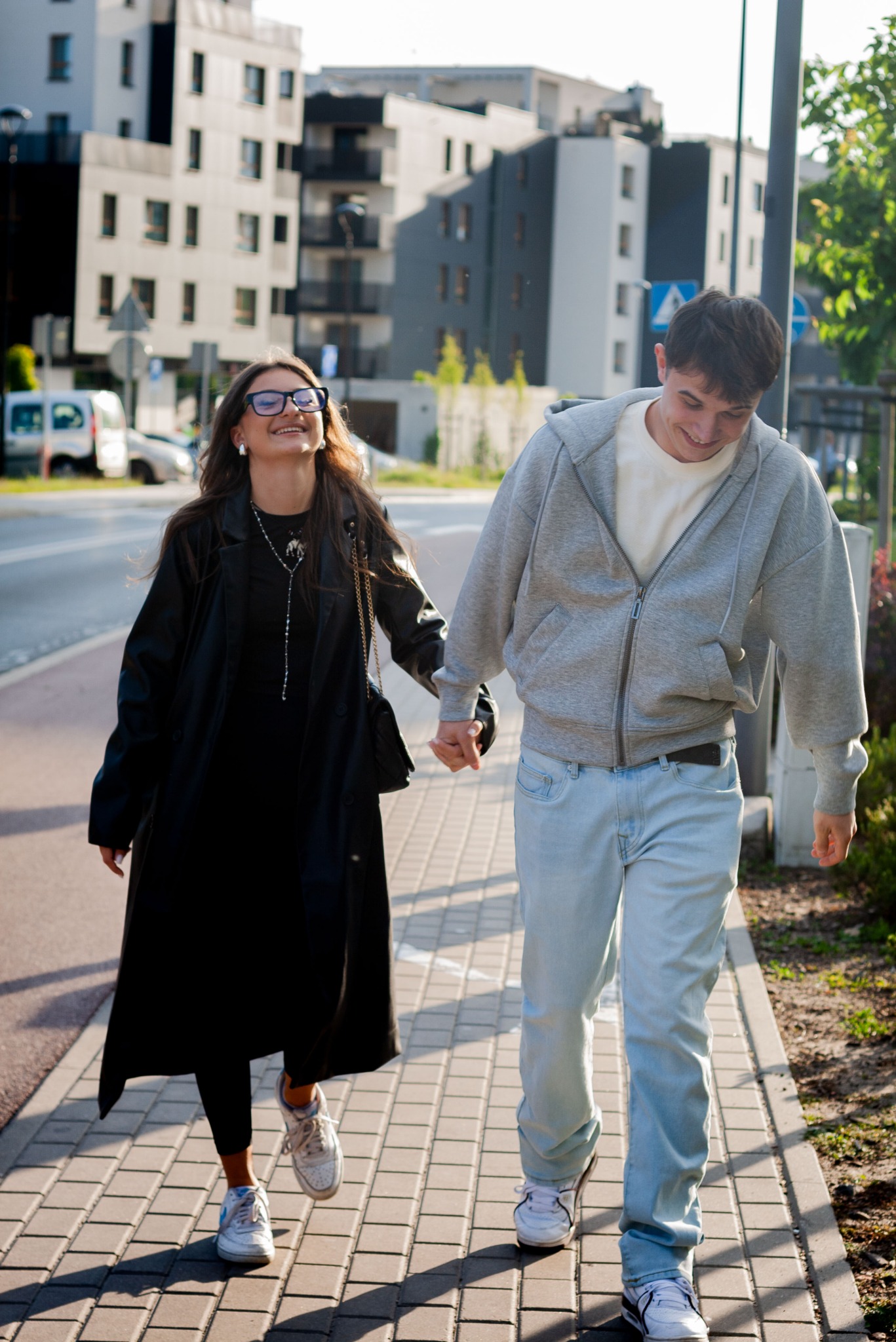 Uśmiechnięta para, ona w czerni i okularach, on w szarej bluzie, idą trzymając się za ręce po chodniku w słoneczny dzień, z budynkami w tle.