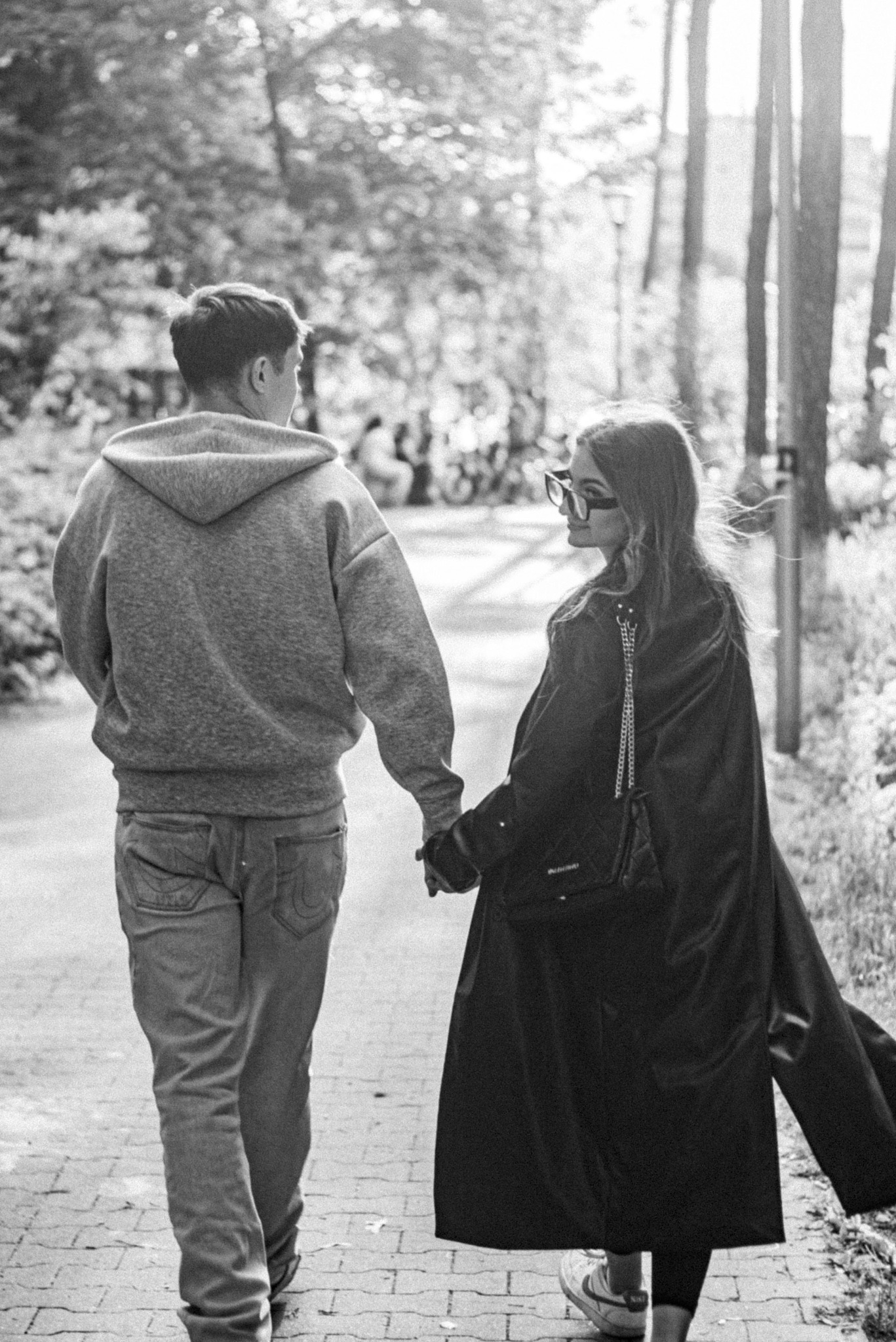 Czarno-białe zdjęcie pary trzymającej się za ręce podczas spaceru w parku, widok z tyłu, z delikatnym światłem słonecznym prześwitującym przez drzewa.