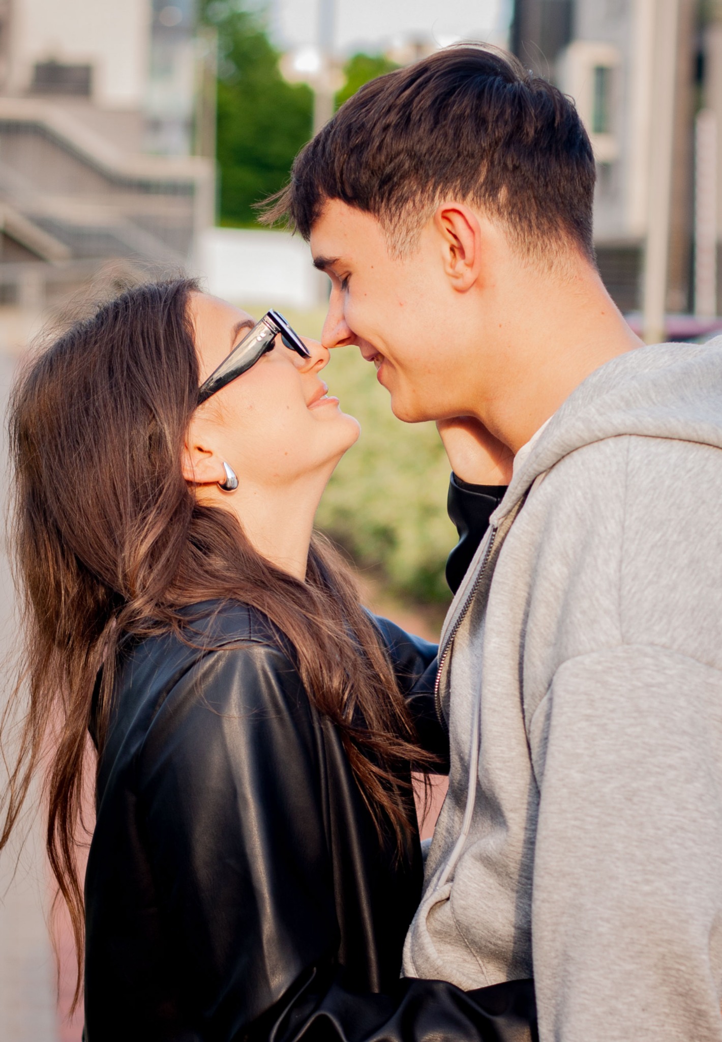 Uśmiechnięta para w zbliżeniu, dotykająca się nosami na tle miejskiej architektury. Dziewczyna w okularach i skórzanej kurtce, chłopak w szarej bluzie z kapturem.