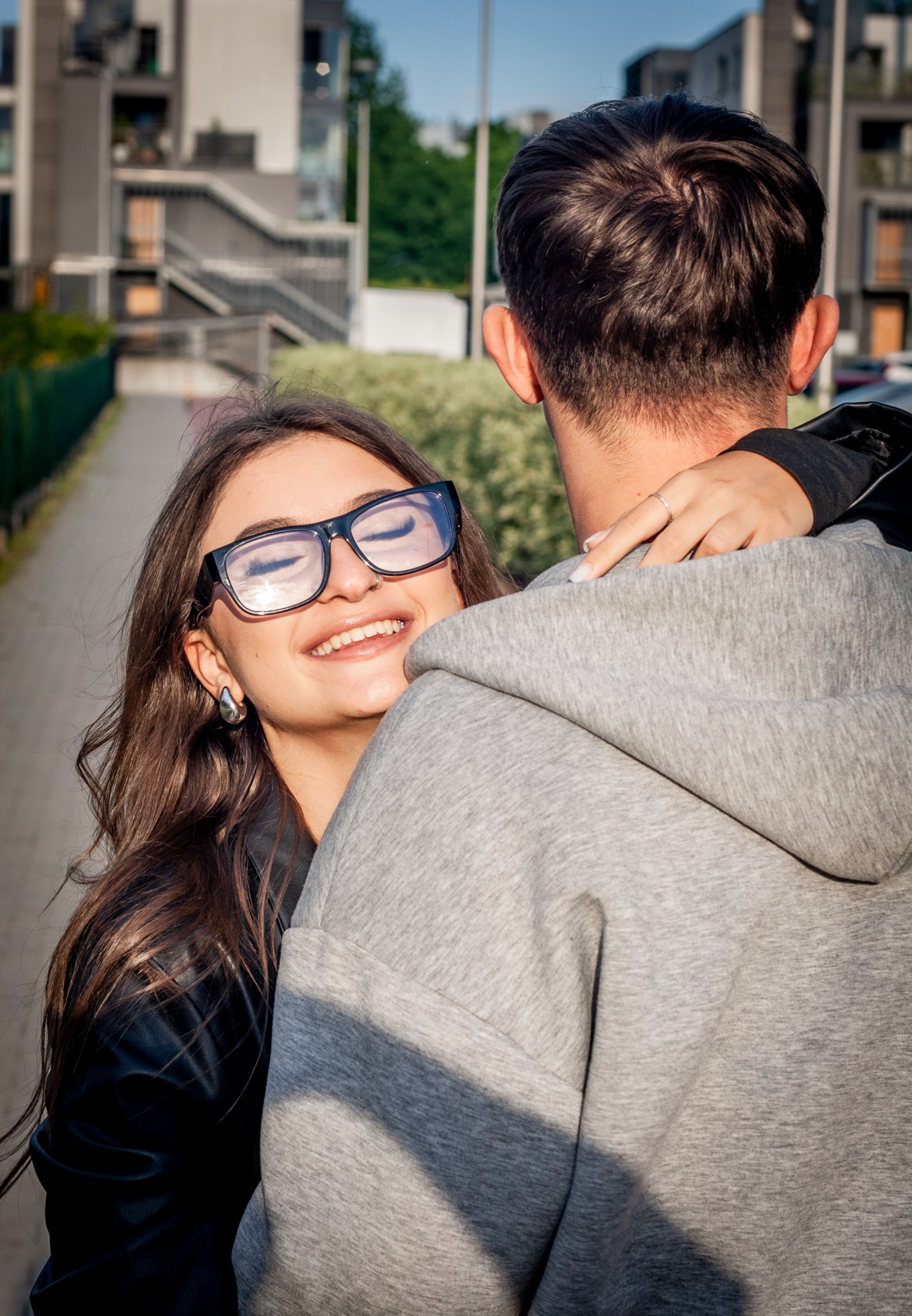 Uśmiechnięta brunetka w okularach obejmuje mężczyznę w szarej bluzie na tle miejskiego pejzażu z budynkami i zielenią. Delikatne światło słoneczne.