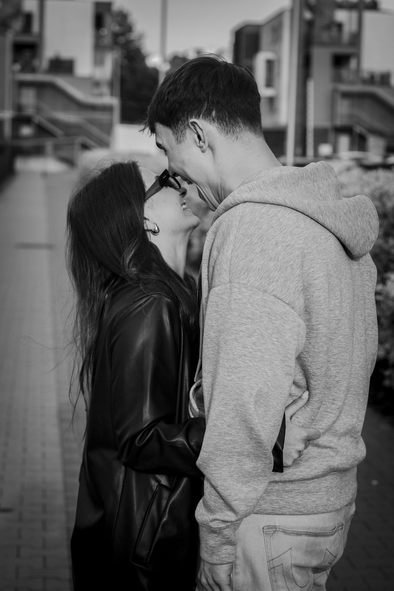 Czarno-białe zdjęcie pary: kobieta w okularach i skórzanej kurtce oraz mężczyzna w bluzie z kapturem, tulący się na tle miejskiego krajobrazu.