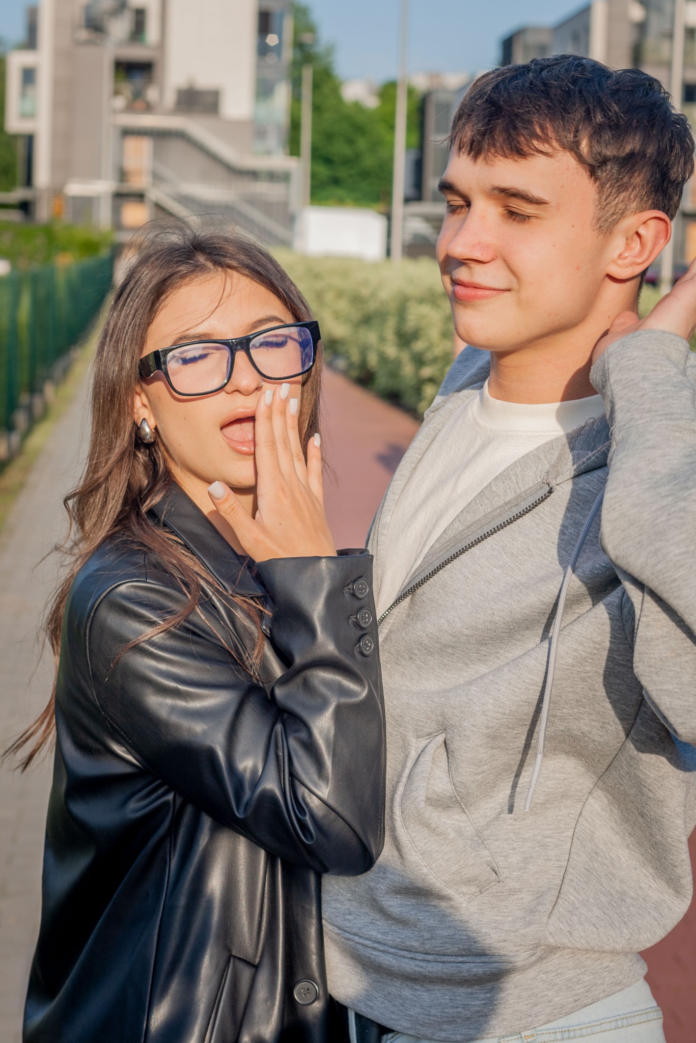 Para młodych ludzi: brunetka w czarnej kurtce i okularach ziewająca, obok niej brunet w szarej bluzie z kapturem, oboje na tle nowoczesnej architektury.