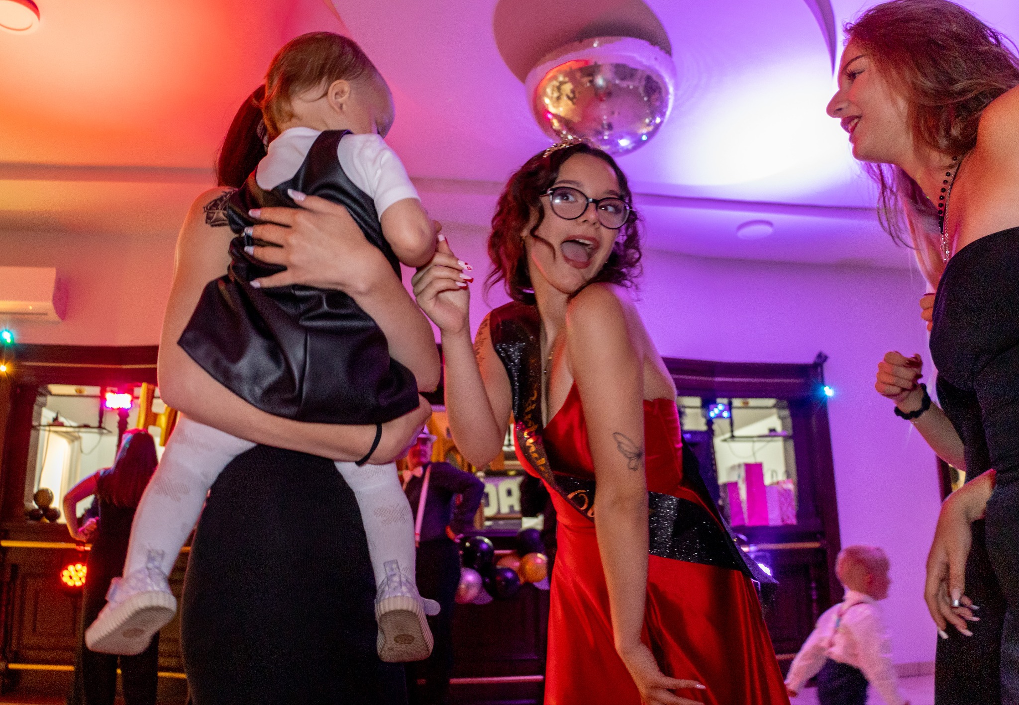 Radosna scena z imprezy: kobieta w czerwonej sukni z szarfą, dziecko na rękach innej kobiety, uśmiechnięta dziewczyna, dyskotekowa kula i dekoracje.
