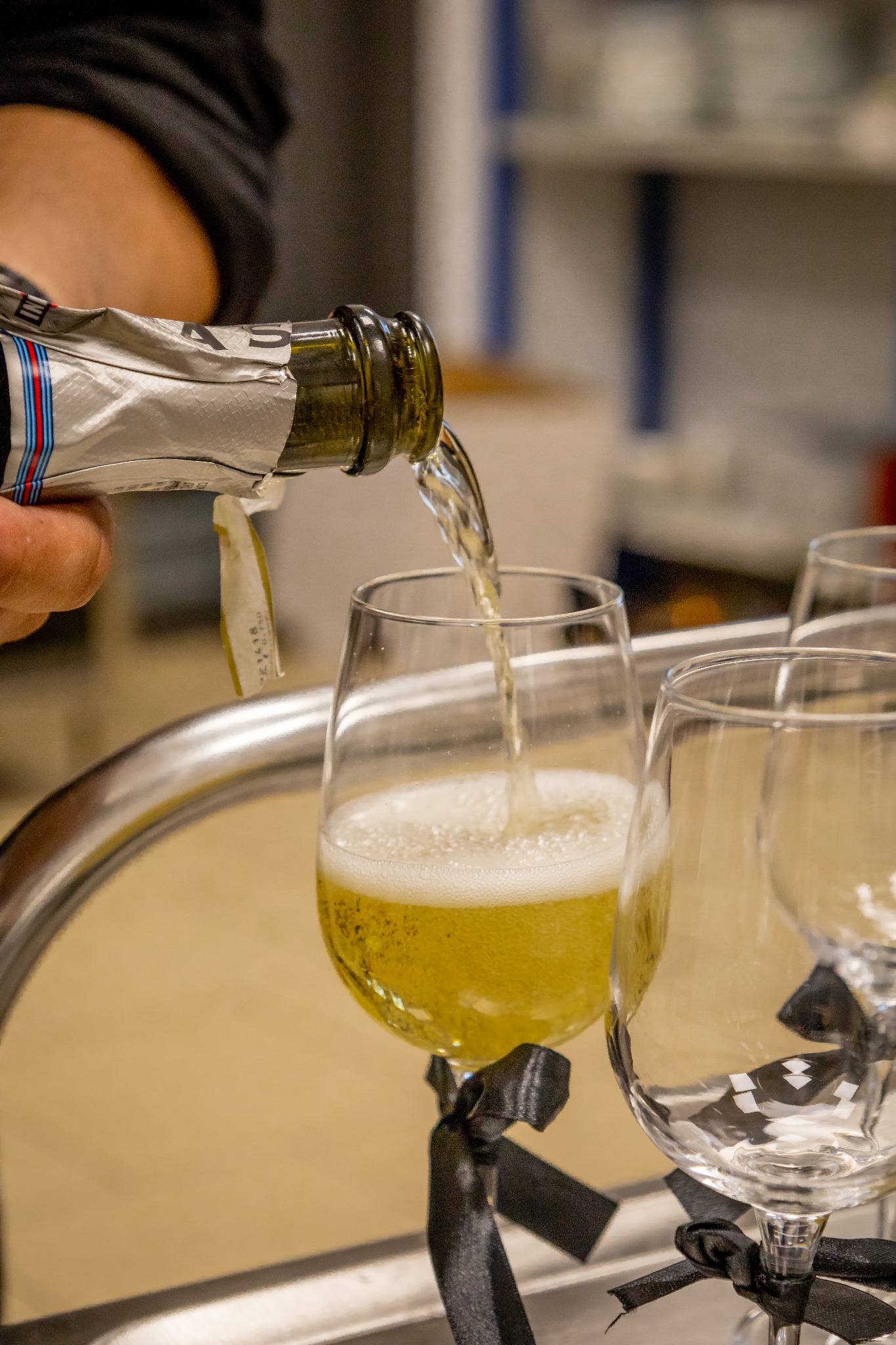 Nalewanie musującego napoju do kieliszka z czarną kokardką. W tle inne kieliszki i butelka. Przygotowania do świętowania.