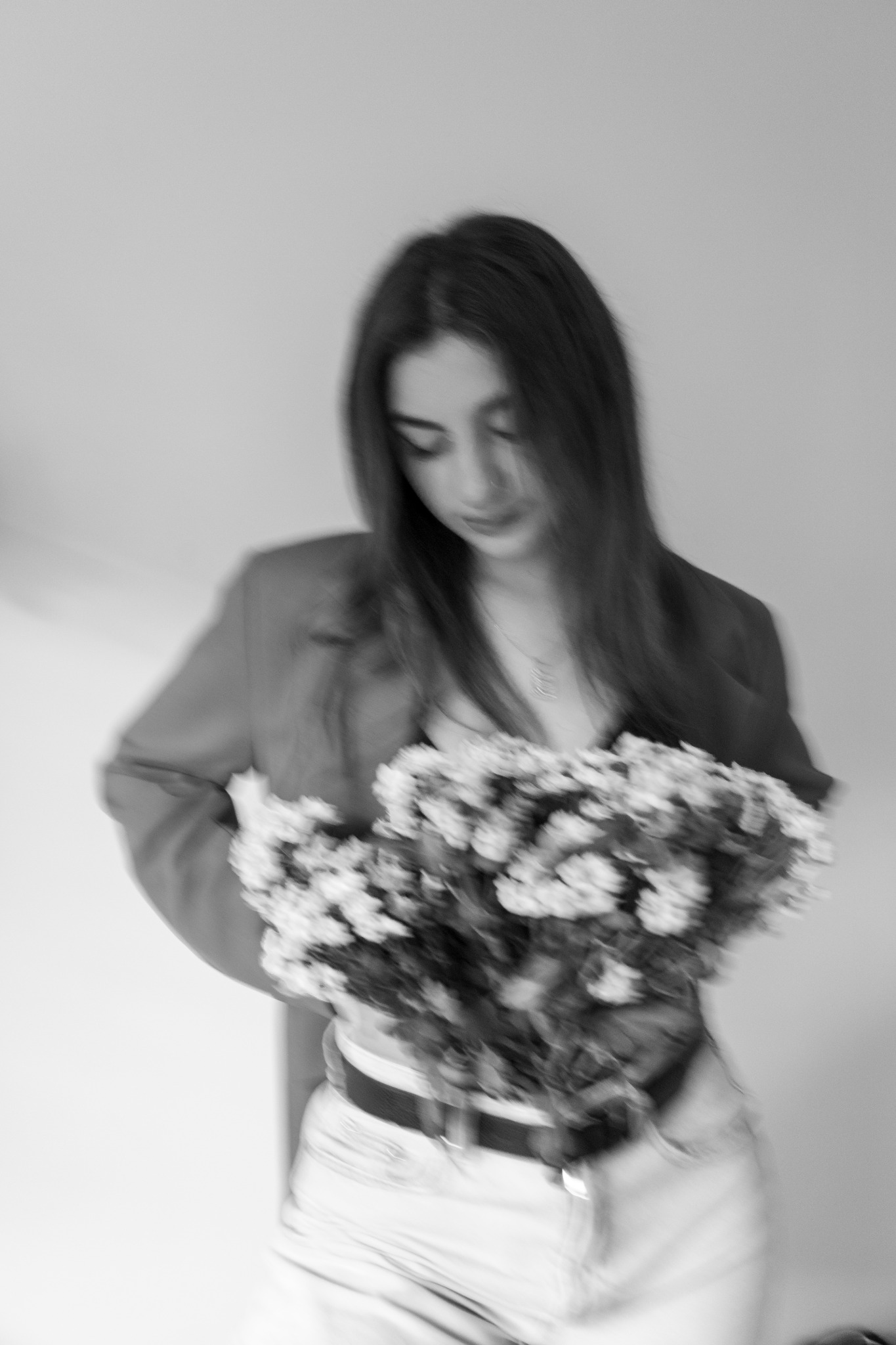 Czarno-białe zdjęcie kobiety z bukietem kwiatów, ubrana w marynarkę i dżinsy. Patrzy w dół na kwiaty, z delikatnym uśmiechem. Rozmycie dodaje dynamiki.