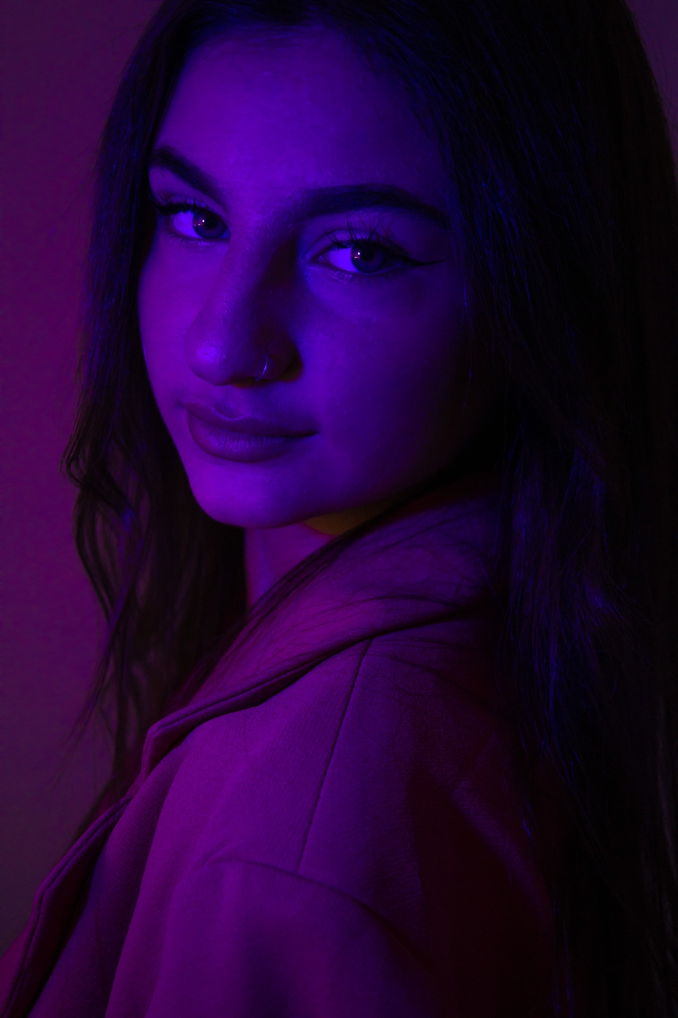 Portret kobiety z kolczykiem w nosie, oświetlonej fioletowym światłem, w marynarce. Ujęcie z bliska, skupione na twarzy i wyrazie oczu.