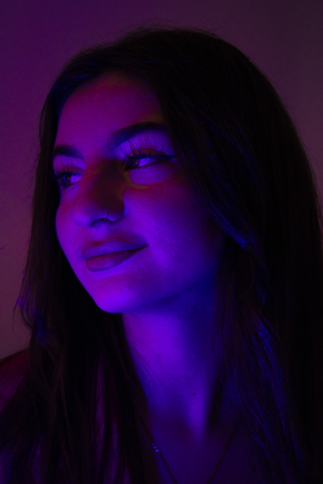 Portret kobiety z kolczykiem w nosie, oświetlonej fioletowo-niebieskim światłem, z wyraźnym makijażem oczu i delikatnym uśmiechem, spoglądającej w lewo.