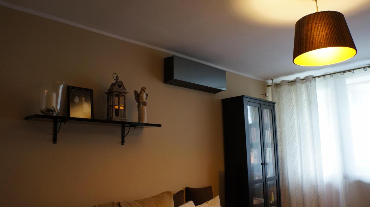 Czarny klimatyzator ścienny w salonie z beżowymi ścianami, półką z dekoracjami i czarną witryną. Ciepłe światło lampy sufitowej.