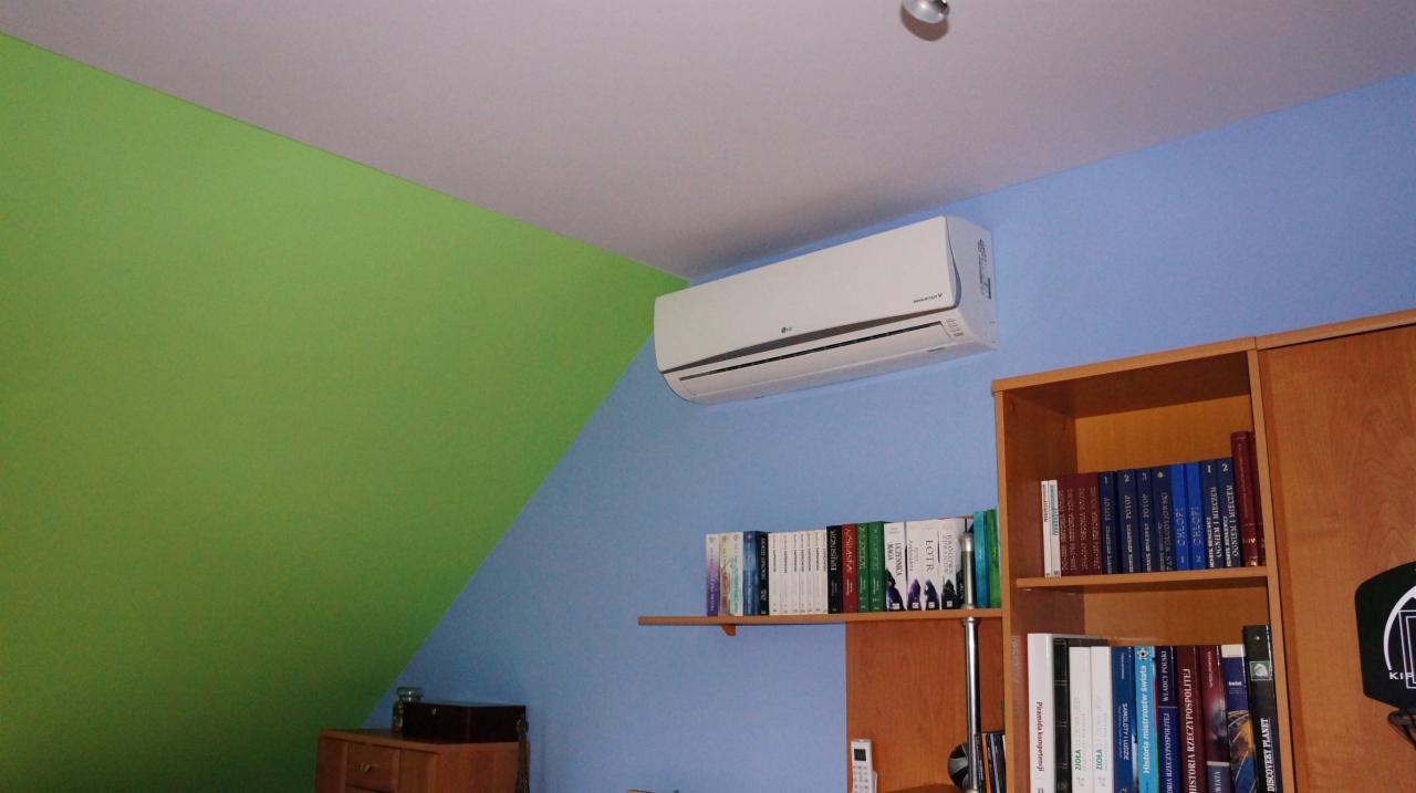 Biały klimatyzator LG zamontowany na błękitnej ścianie w pokoju z zielonym akcentem, obok drewnianego regału z książkami. Widoczny pilot i fragment komody.