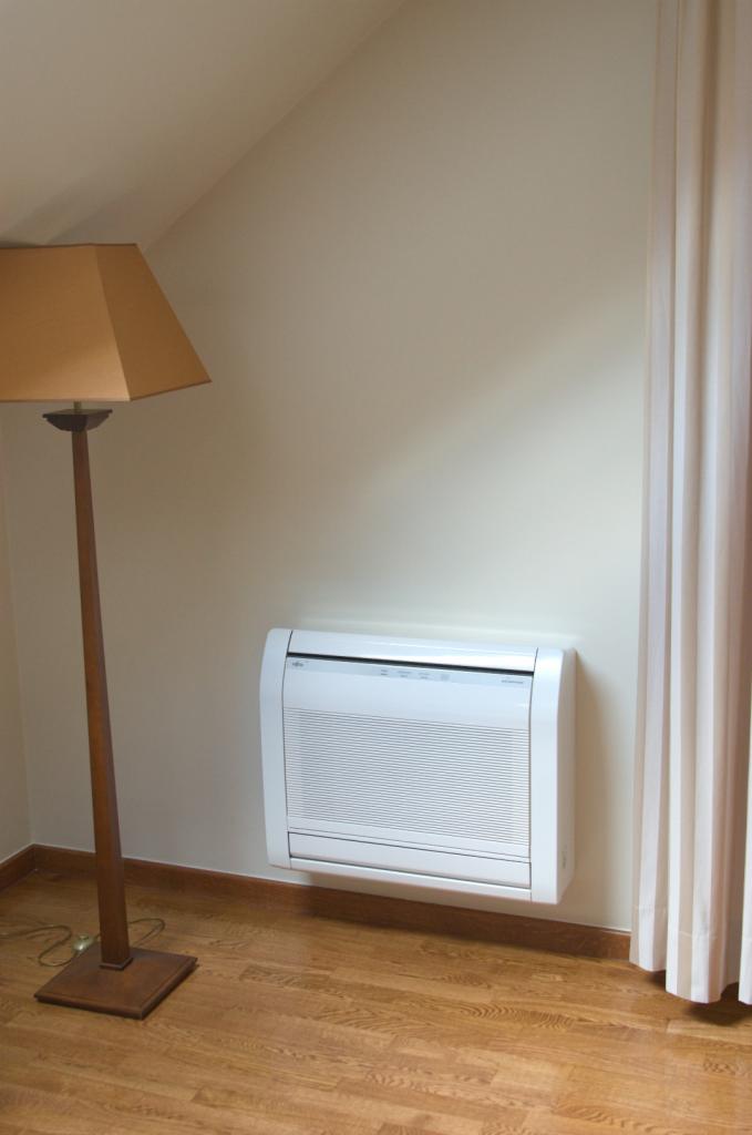 Biała, naścienna jednostka klimatyzacyjna Fujitsu w pokoju z drewnianą podłogą, obok lampy stojącej i zasłony. Neutralne kolory ścian.