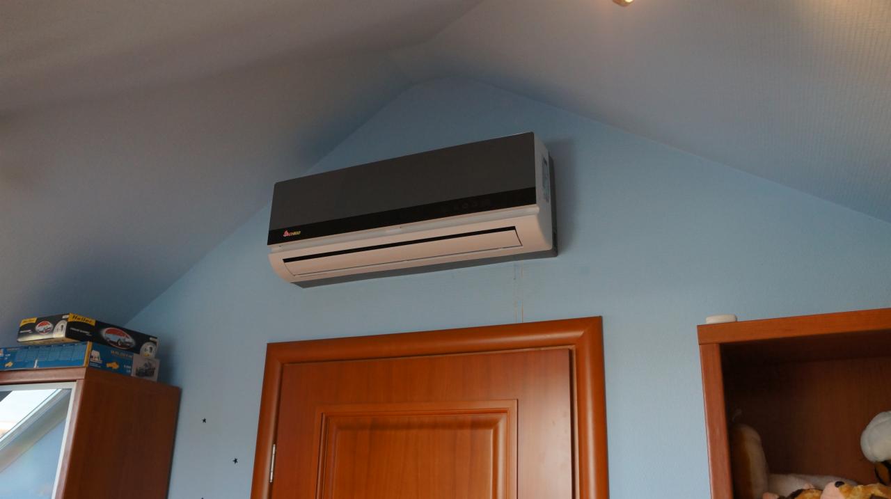 Szara klimatyzacja ścienna zamontowana nad brązowymi drzwiami w pokoju z niebieskimi ścianami i skośnym sufitem. Widoczny fragment mebli.