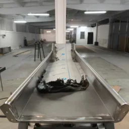 Metalowa konstrukcja z blachy, prawdopodobnie element linii produkcyjnej, z resztkami folii ochronnej, w hali przemysłowej.