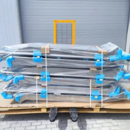 Stalowe konstrukcje zabezpieczone folią i pianką, ułożone na palecie przed szarym budynkiem. Elementy ślusarki w transporcie.