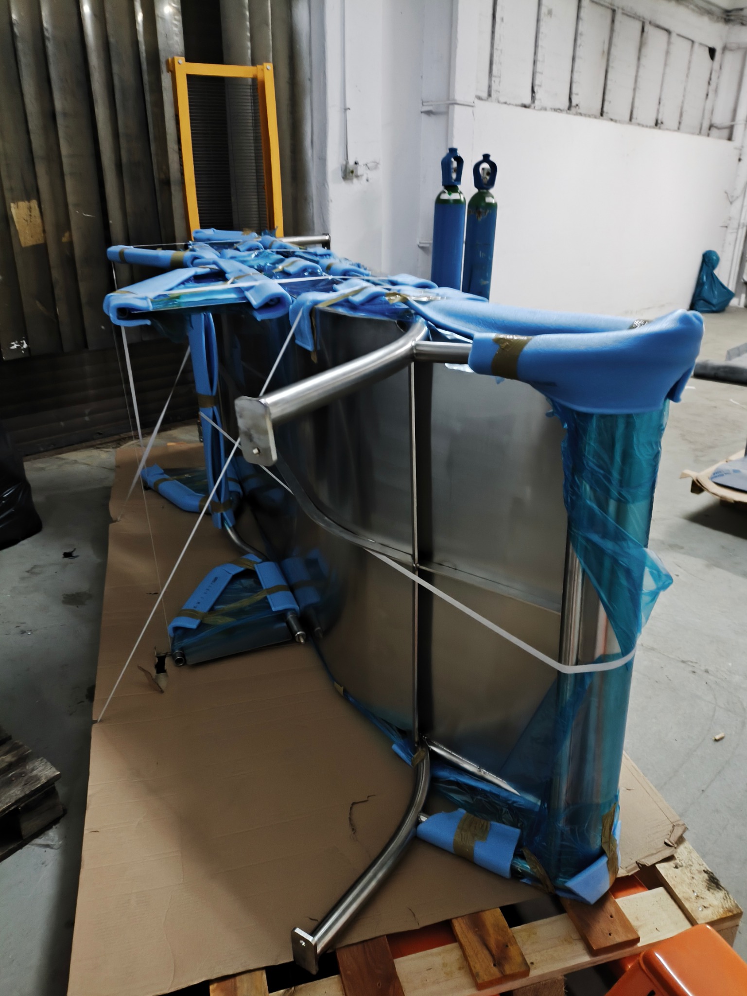 Element konstrukcyjny ze stali nierdzewnej, zabezpieczony niebieską folią ochronną i pianką, umieszczony na palecie z tektury falistej w magazynie, obok butle z gazem.