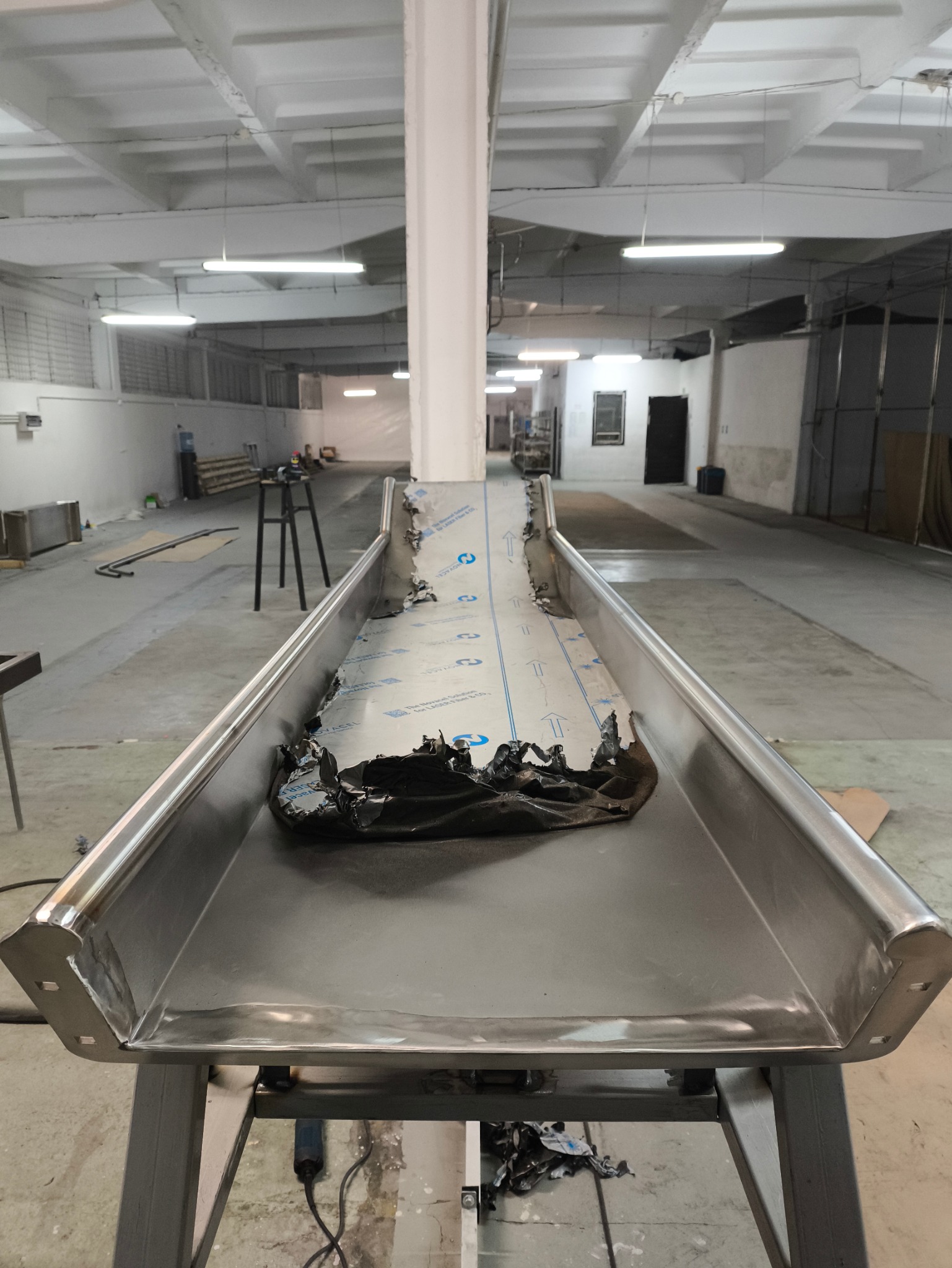 Metalowa konstrukcja z blachy, prawdopodobnie element linii produkcyjnej, z resztkami folii ochronnej, w hali przemysłowej.