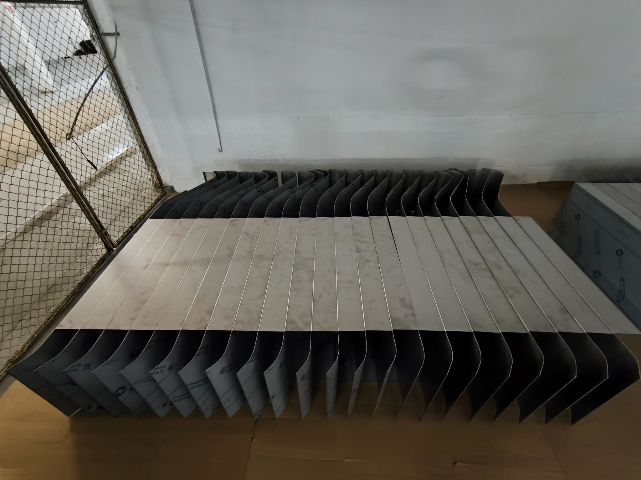 Stos wyciętych, giętych elementów ze stali nierdzewnej, prawdopodobnie do balustrady, z widoczną folią ochronną na niektórych elementach.