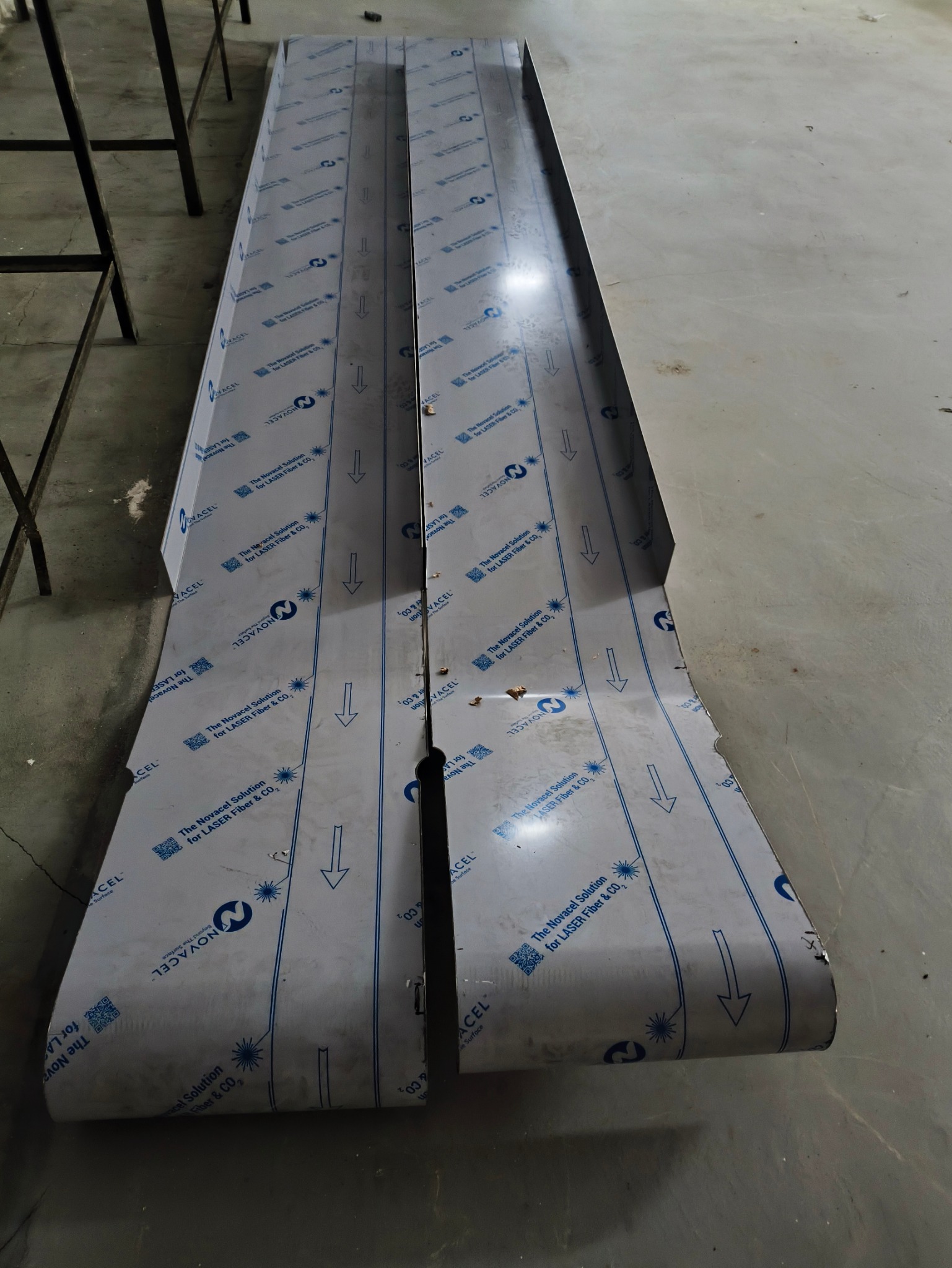 Dwa elementy z blachy nierdzewnej pokryte folią ochronną z nadrukiem, leżące na podłodze warsztatu.