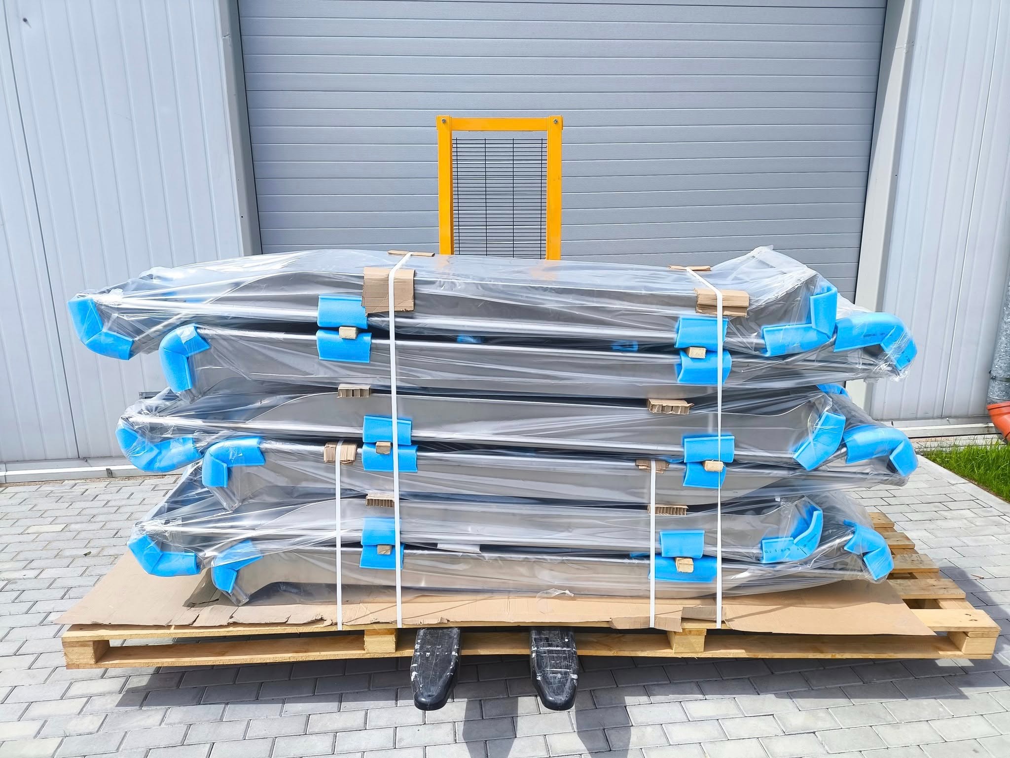 Stalowe konstrukcje zabezpieczone folią i pianką, ułożone na palecie przed szarym budynkiem. Elementy ślusarki w transporcie.