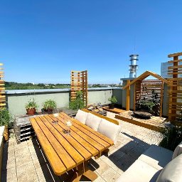 HomeSpace Biuro nieruchomości Rzeszów - Słoneczny taras na dachu budynku z drewnianymi meblami ogrodowymi, pergolą i roślinami w doniczkach, widok na miasto w tle.