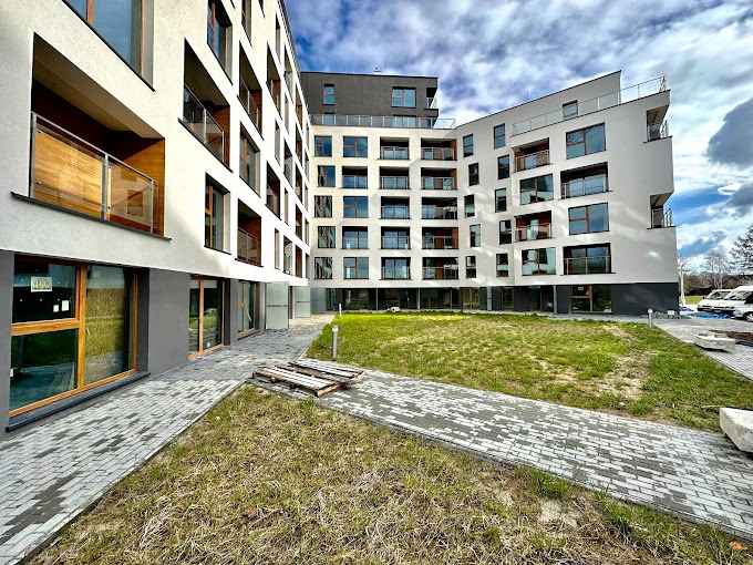 Nowoczesny budynek mieszkalny z jasną elewacją i balkonami z drewnianymi panelami, widok z poziomu chodnika z kostki brukowej na trawnik przed budynkiem, błękitne niebo z chmurami w tle.