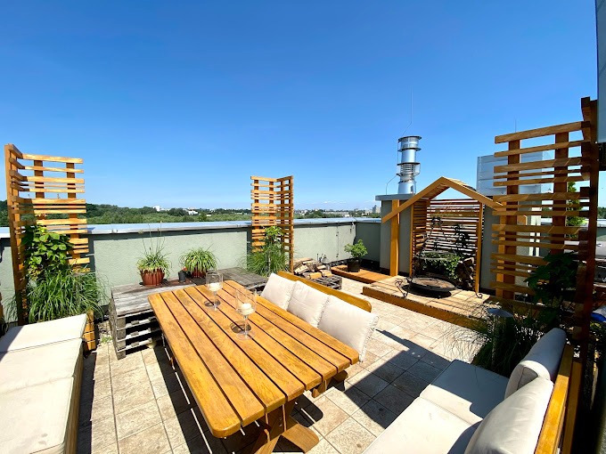 Słoneczny taras na dachu budynku z drewnianymi meblami ogrodowymi, pergolą i roślinami w doniczkach, widok na miasto w tle.