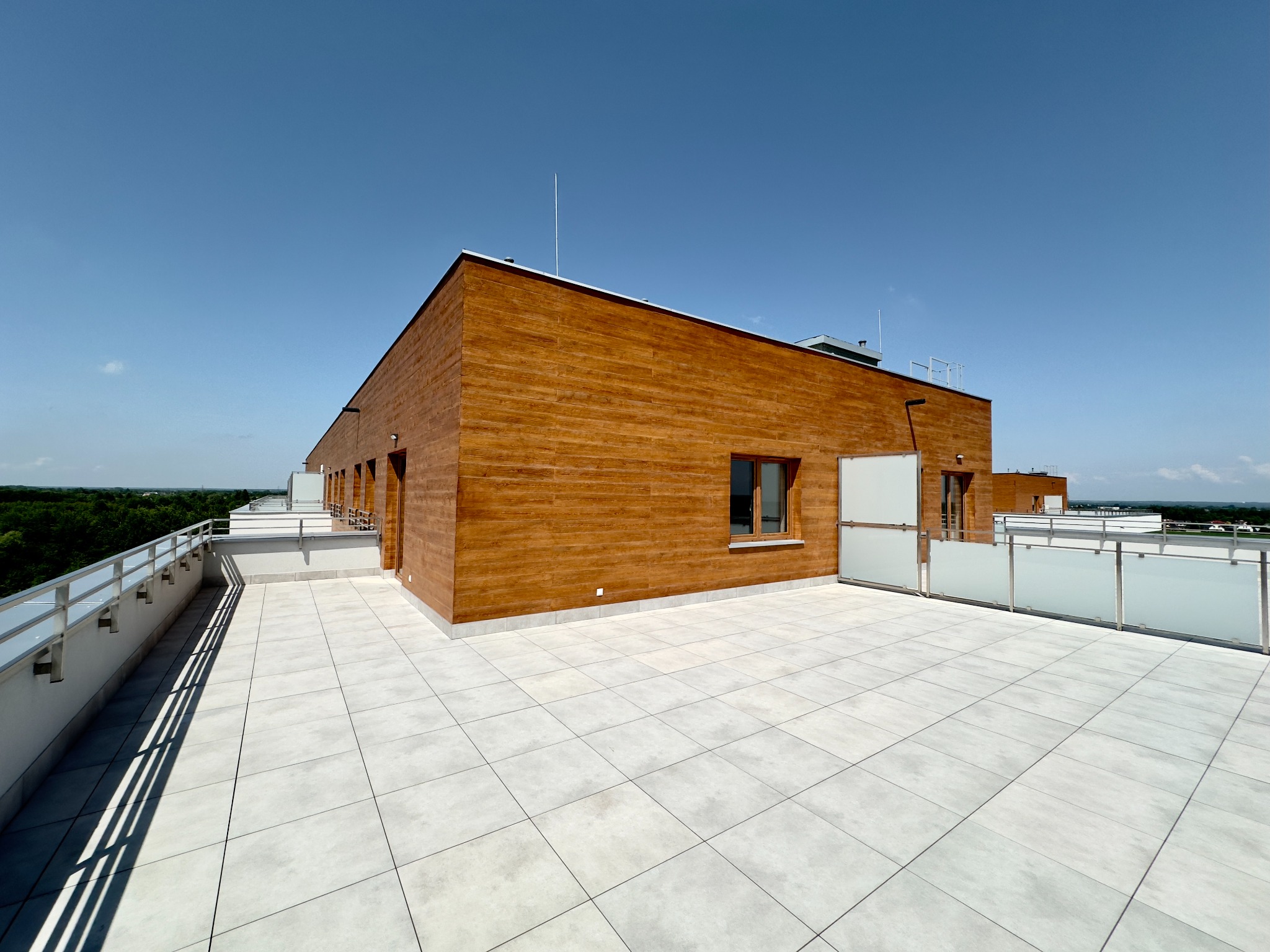 Nowoczesny budynek z elewacją z drewnopodobnych paneli, widoczny z przestronnego tarasu wyłożonego jasnymi płytkami, barierki ze stali nierdzewnej z matowym szkłem, błękitne niebo w tle.