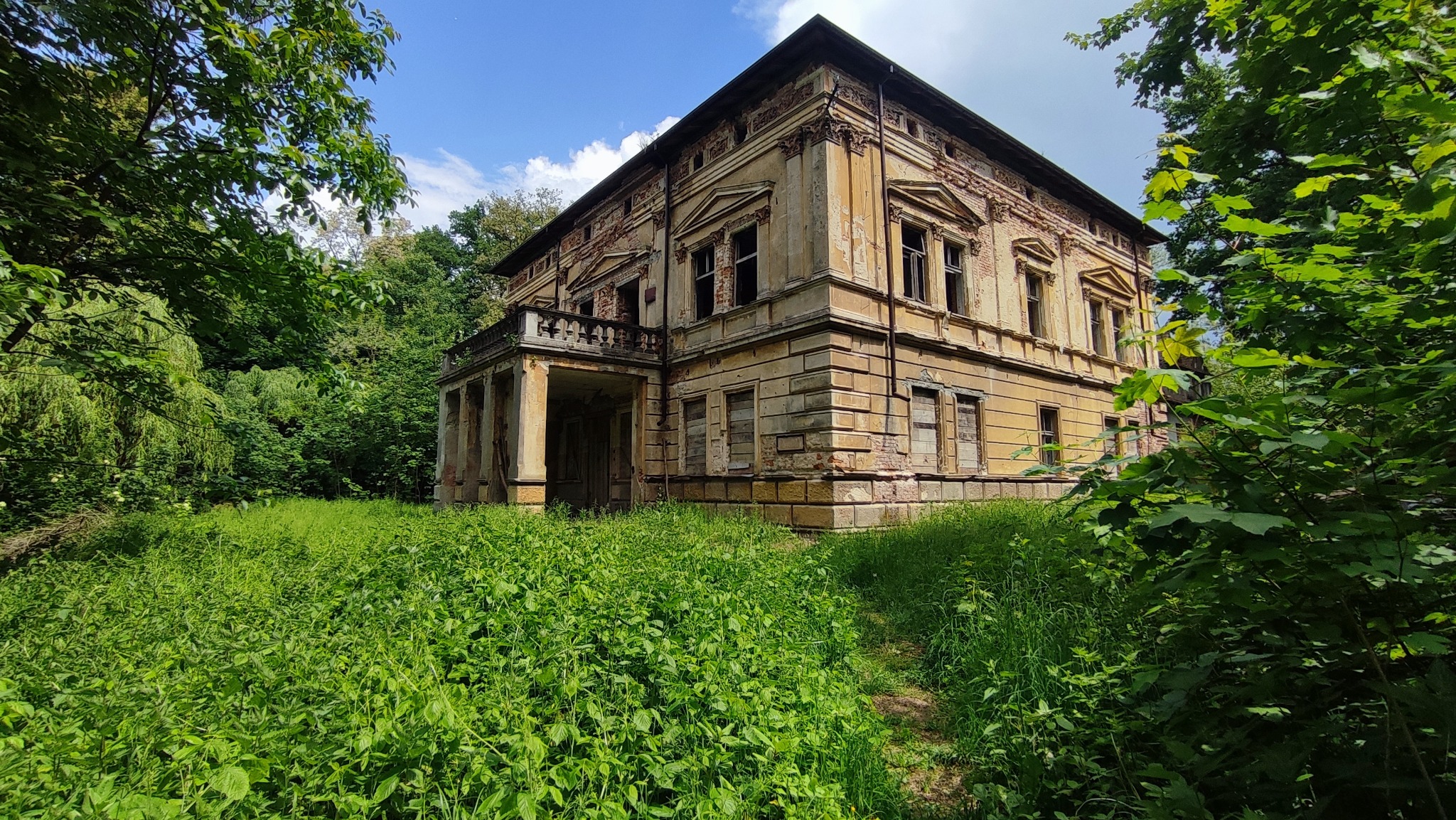 Zrujnowany, dwupiętrowy budynek z kolumnami i balkonem, porośnięty bujną zielenią, z widocznymi śladami zaniedbania i uszkodzeń elewacji.