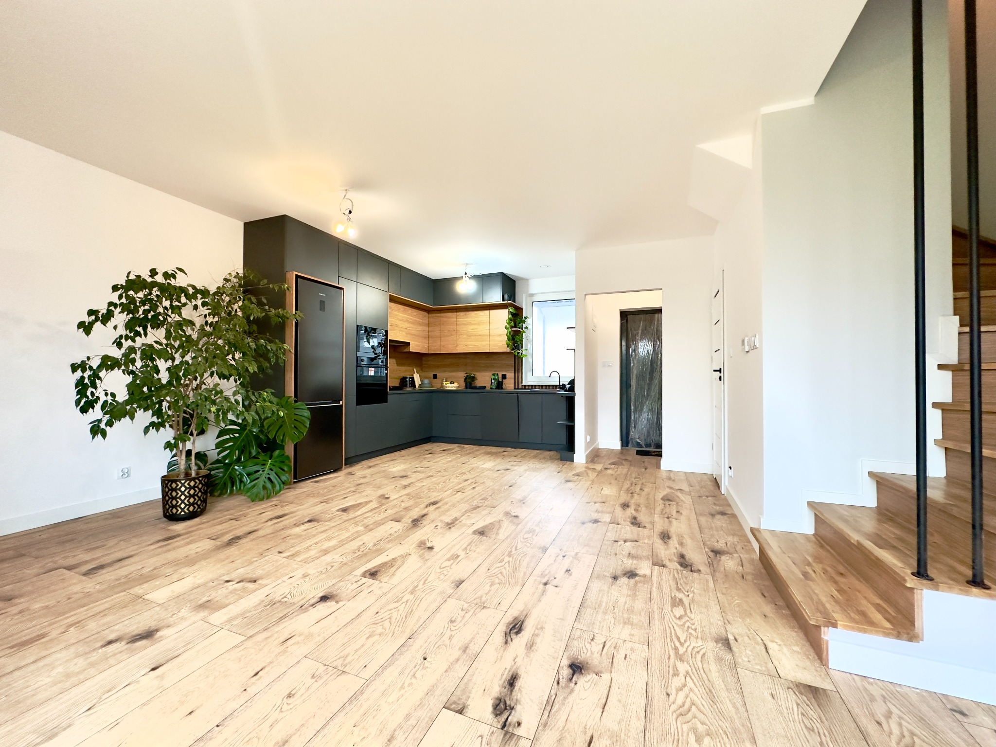 Przestronne wnętrze domu z parkietem, ciemną zabudową kuchenną, schodami z drewnianymi stopniami i zielonymi roślinami doniczkowymi