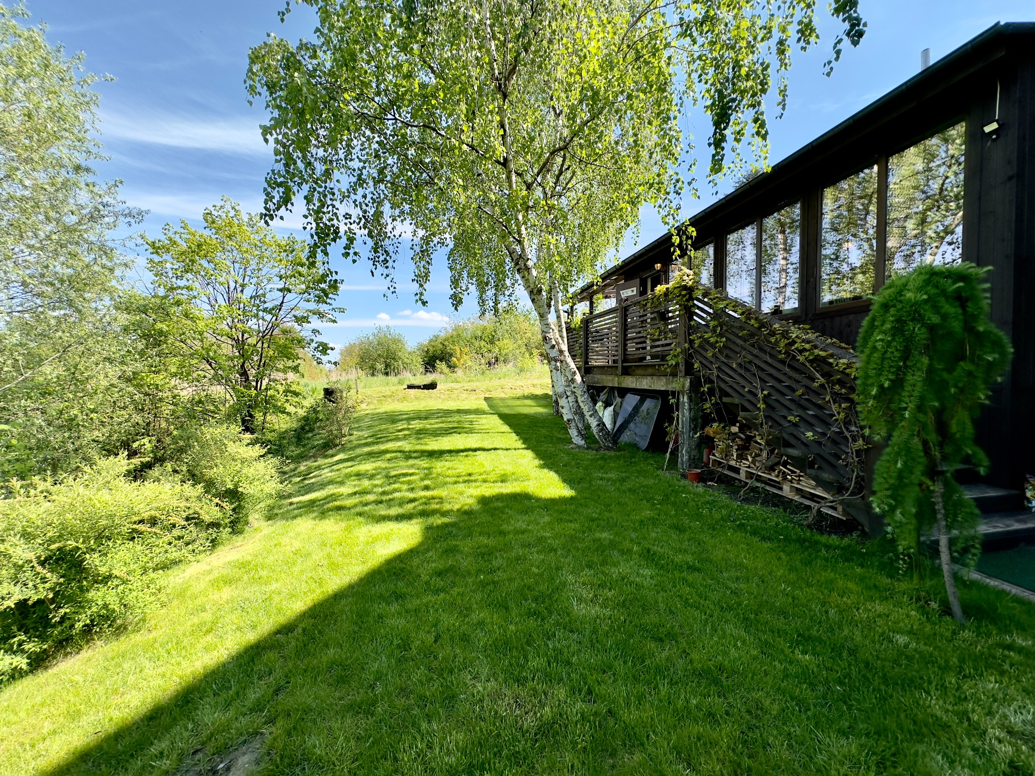 Nowoczesny, czarny dom z dużymi oknami i drewnianym balkonem, otoczony zielonym trawnikiem i drzewami, w słoneczny dzień.