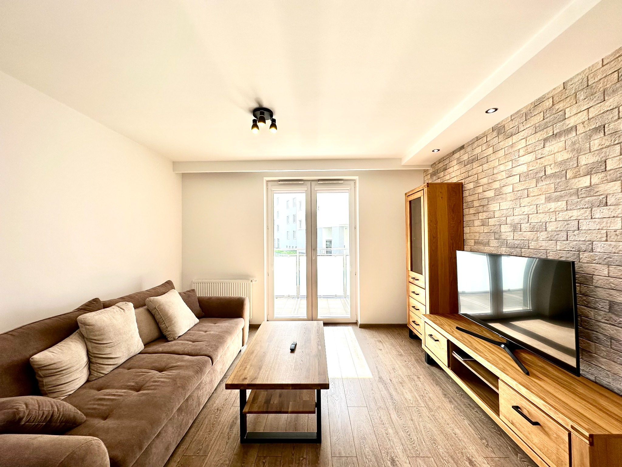 Jasne wnętrze salonu z brązową sofą, drewnianym stolikiem kawowym z pilotem, szafką RTV i telewizorem na tle ściany z dekoracyjnej cegły.