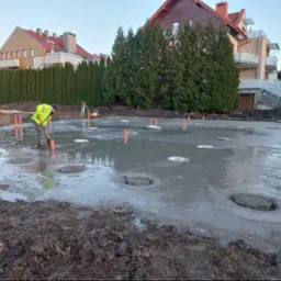 Wylewka fundamentu z widocznymi otworami na słupy, pracownik w odblaskowej kamizelce wyrównuje powierzchnię, w tle zabudowa jednorodzinna i rząd tuj.