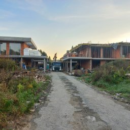 VegasBUD - Wczesny etap budowy dwóch domów jednorodzinnych z czerwonej cegły, widok z drogi dojazdowej, z widocznym samochodem ciężarowym z dźwigiem w tle.