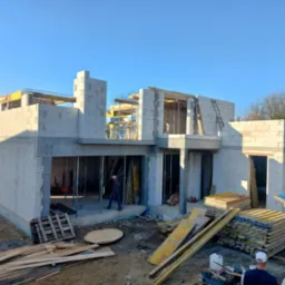 Wznoszenie ścian parterowego budynku z betonu komórkowego, widoczne szalunki stropu, zbrojenia i praca dwóch robotników budowlanych.