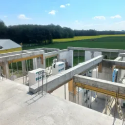 Surowy stan budynku z betonu komórkowego H+H w trakcie budowy, widok z góry na konstrukcję zbrojoną, w tle zielone pola i las, błękitne niebo.