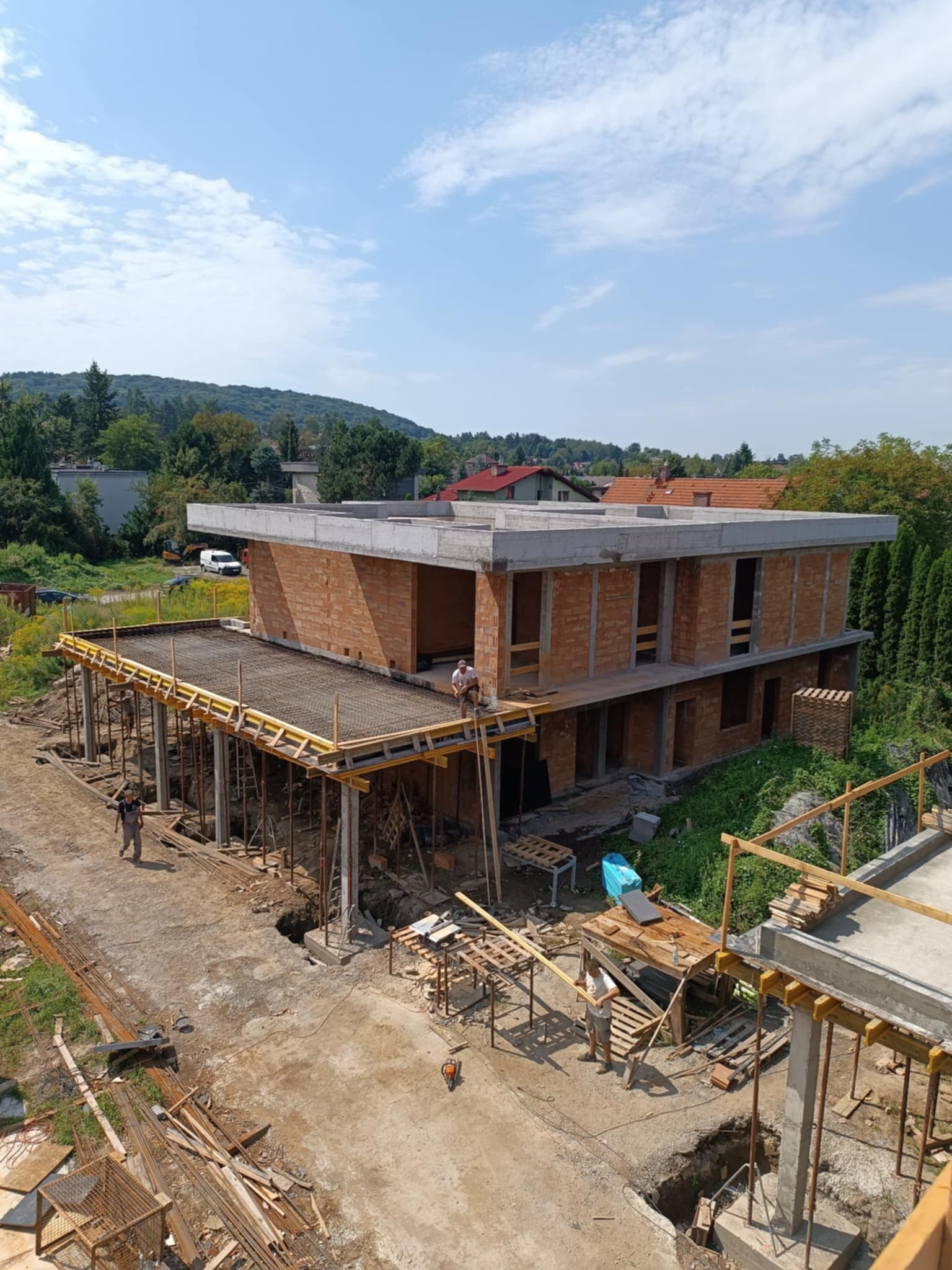 Dwukondygnacyjny budynek w stanie surowym otwartym, z widocznymi elementami konstrukcji żelbetowej i murowanymi ścianami z cegły, na pierwszym planie szalunek stropu zbrojonego, robotnicy przy...