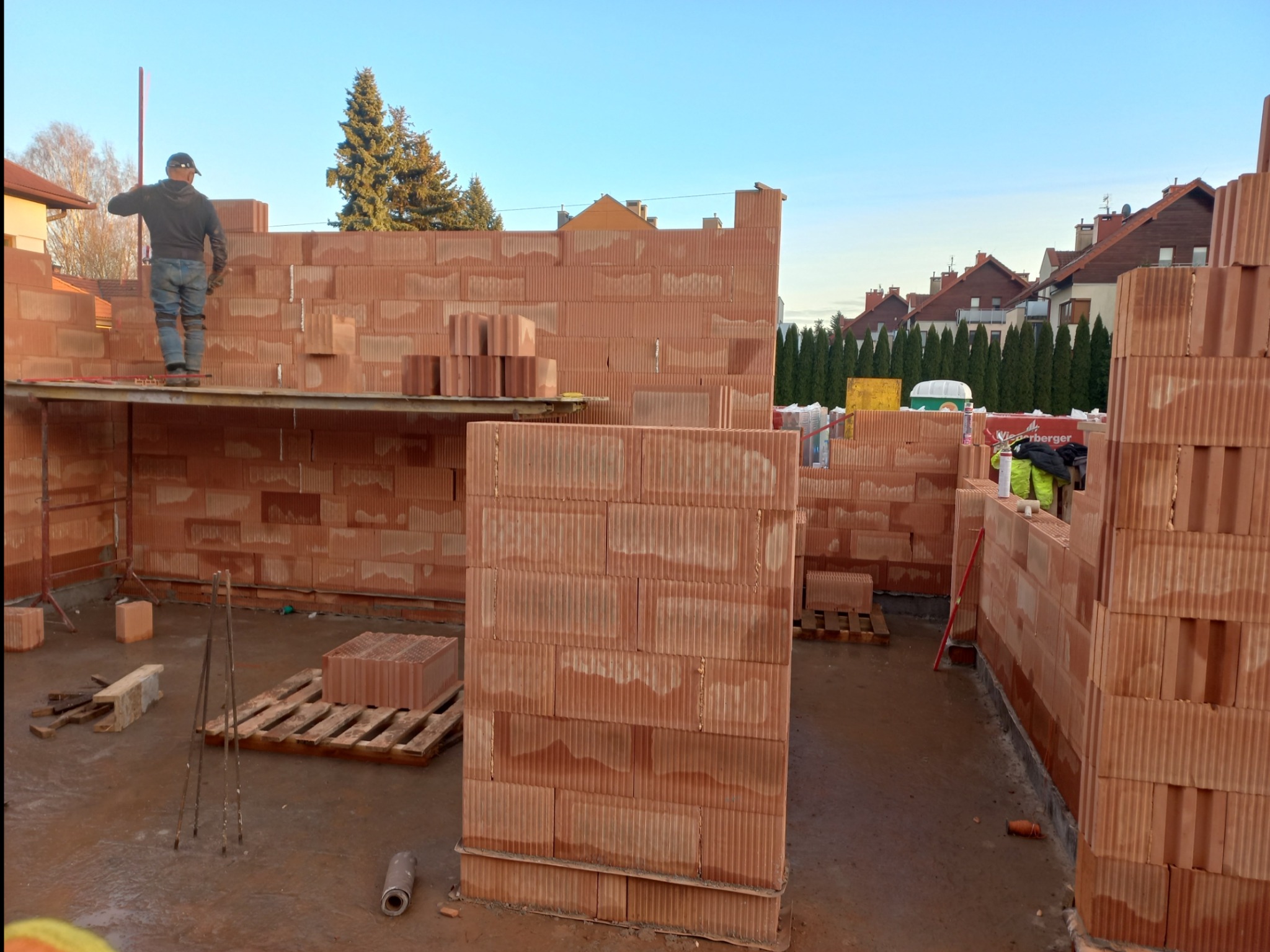 Wznoszenie ścian zewnętrznych z pustaków ceramicznych na placu budowy, widoczny pracownik na rusztowaniu, palety z materiałem i narzędzia murarskie.