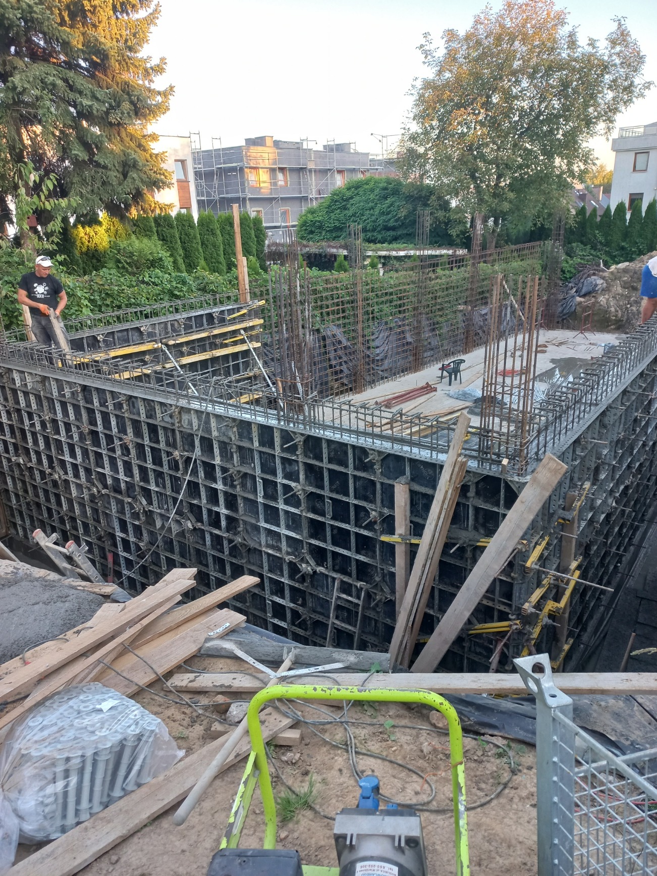 Wykop pod fundamenty z szalunkiem systemowym i zbrojeniem, widoczny pracownik w tle oraz zagęszczarka wibracyjna na pierwszym planie.