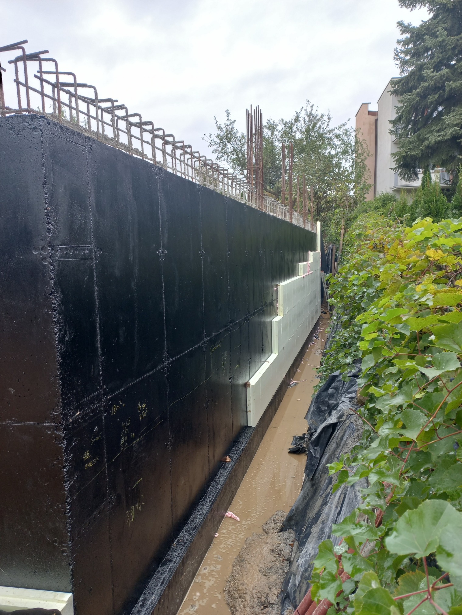 Izolacja pionowa fundamentu z czarną powłoką hydroizolacyjną i białymi płytami styropianu, widoczne zbrojenia wystające ponad poziom gruntu, teren po deszczu.