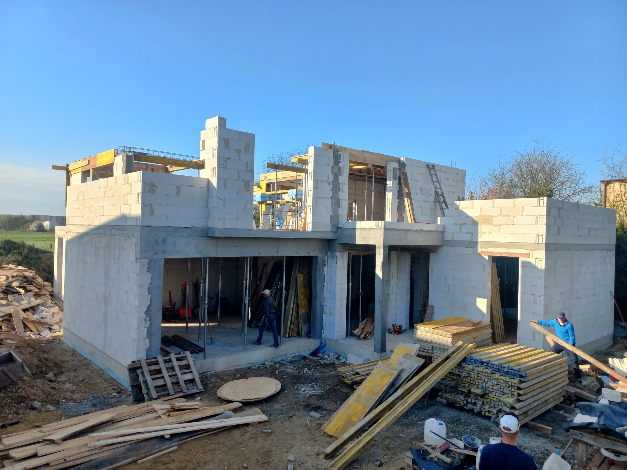 Wznoszenie ścian parterowego budynku z betonu komórkowego, widoczne szalunki stropu, zbrojenia i praca dwóch robotników budowlanych.