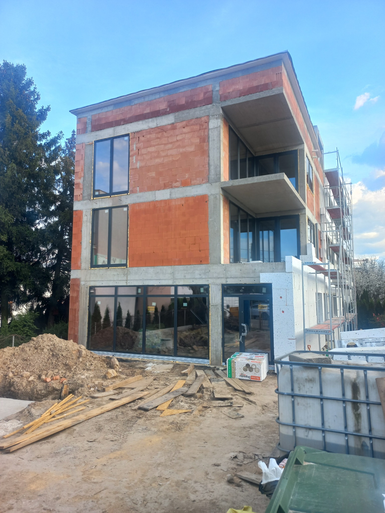 Trzykondygnacyjny budynek w trakcie budowy z czerwonej cegły i betonu, z widocznymi oknami i balkonami, otoczony materiałami budowlanymi i rusztowaniami.