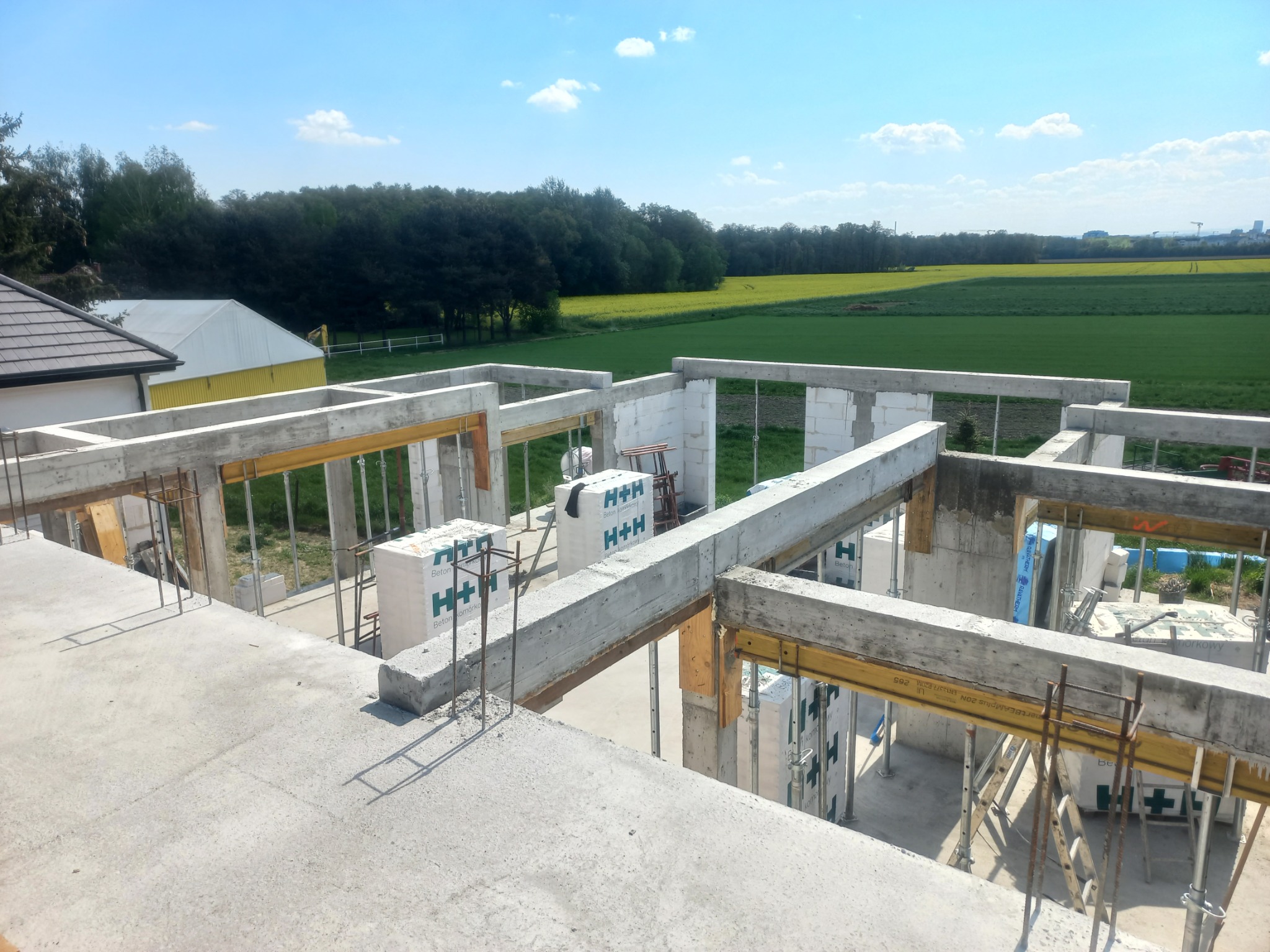 Surowy stan budynku z betonu komórkowego H+H w trakcie budowy, widok z góry na konstrukcję zbrojoną, w tle zielone pola i las, błękitne niebo.